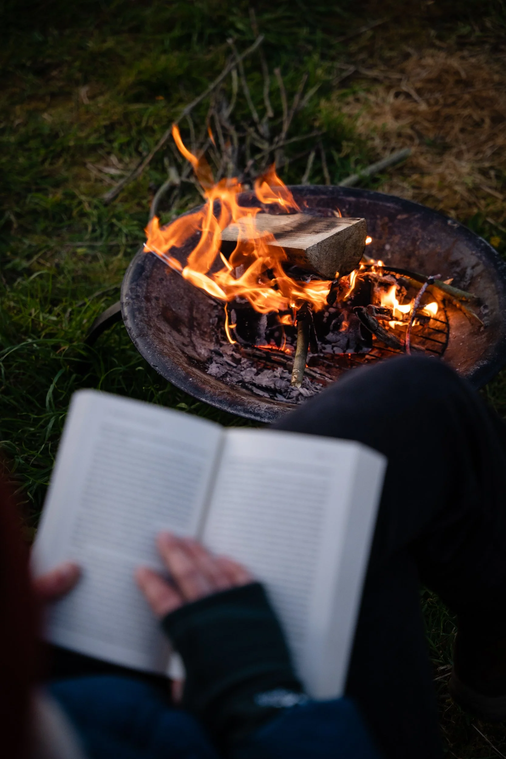 Person reading a book near a fire pit with burning wood outdoors.