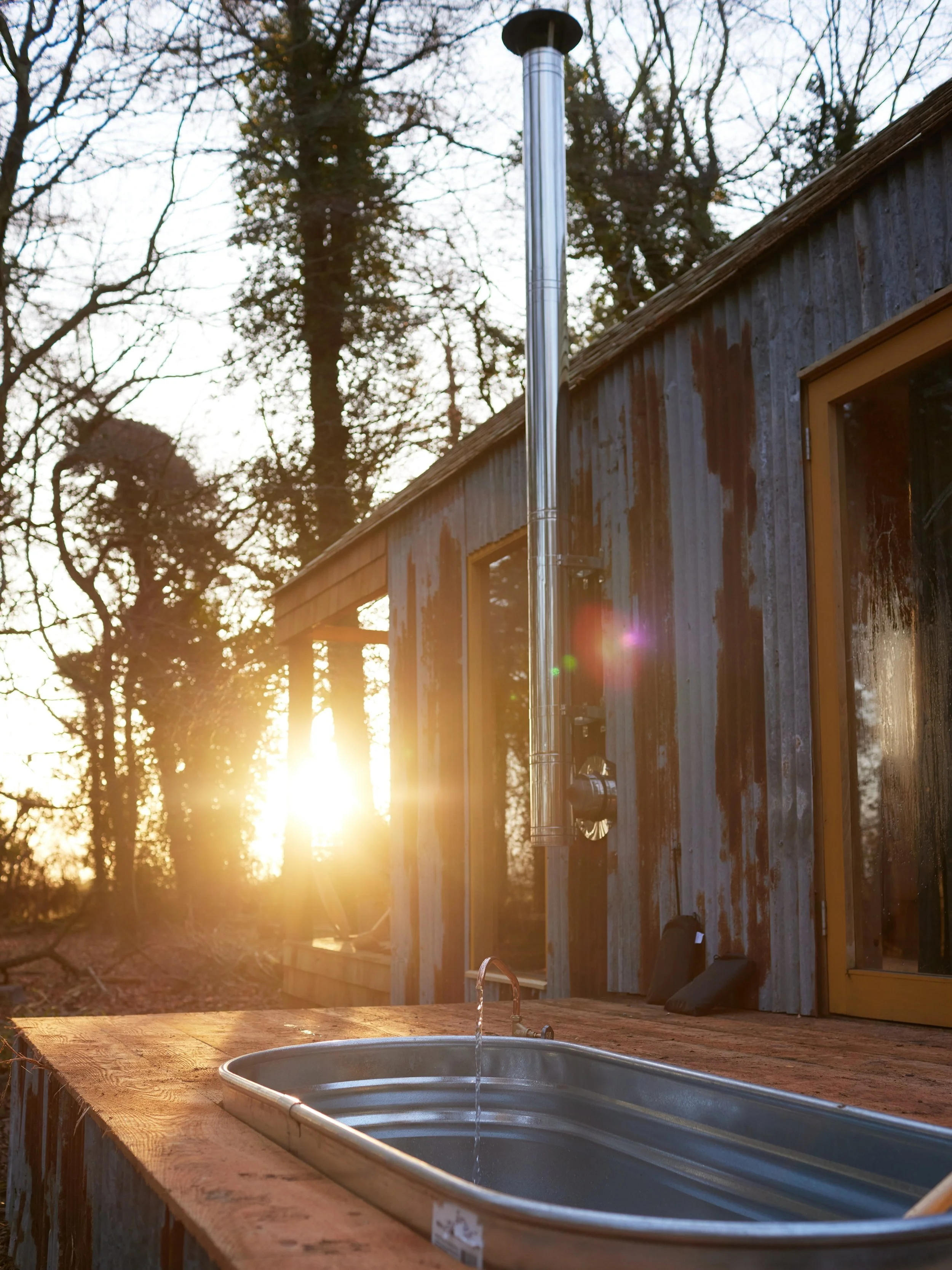 Outdoor sink with running water on a wooden counter beside a rustic metal building, surrounded by trees, during a sunset