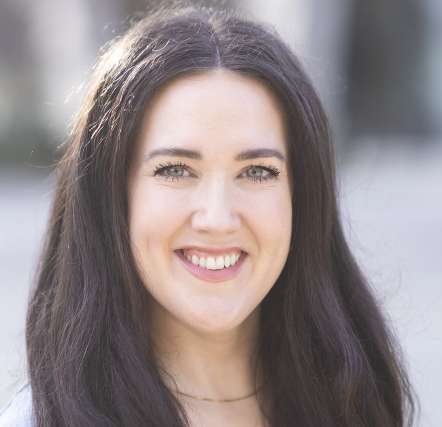Portrait of Laura Berringer, an expert in neurodiversity, particular autism and behavior.  It is a close-up portrait. She has long dark hair, light-colored eyes, smiling showing her teeth, outdoors with blurred background.