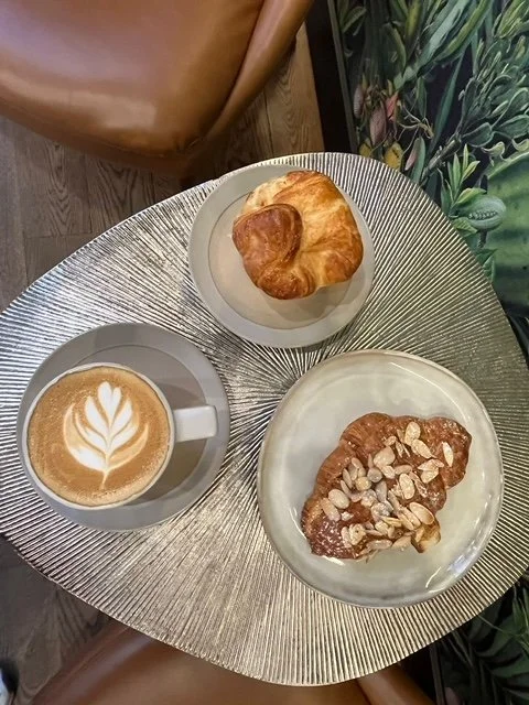 A silver textured round table with a latte coffee with latte art, a croissant on a plate, and a slice of toast topped with sunflower seeds and nuts.
