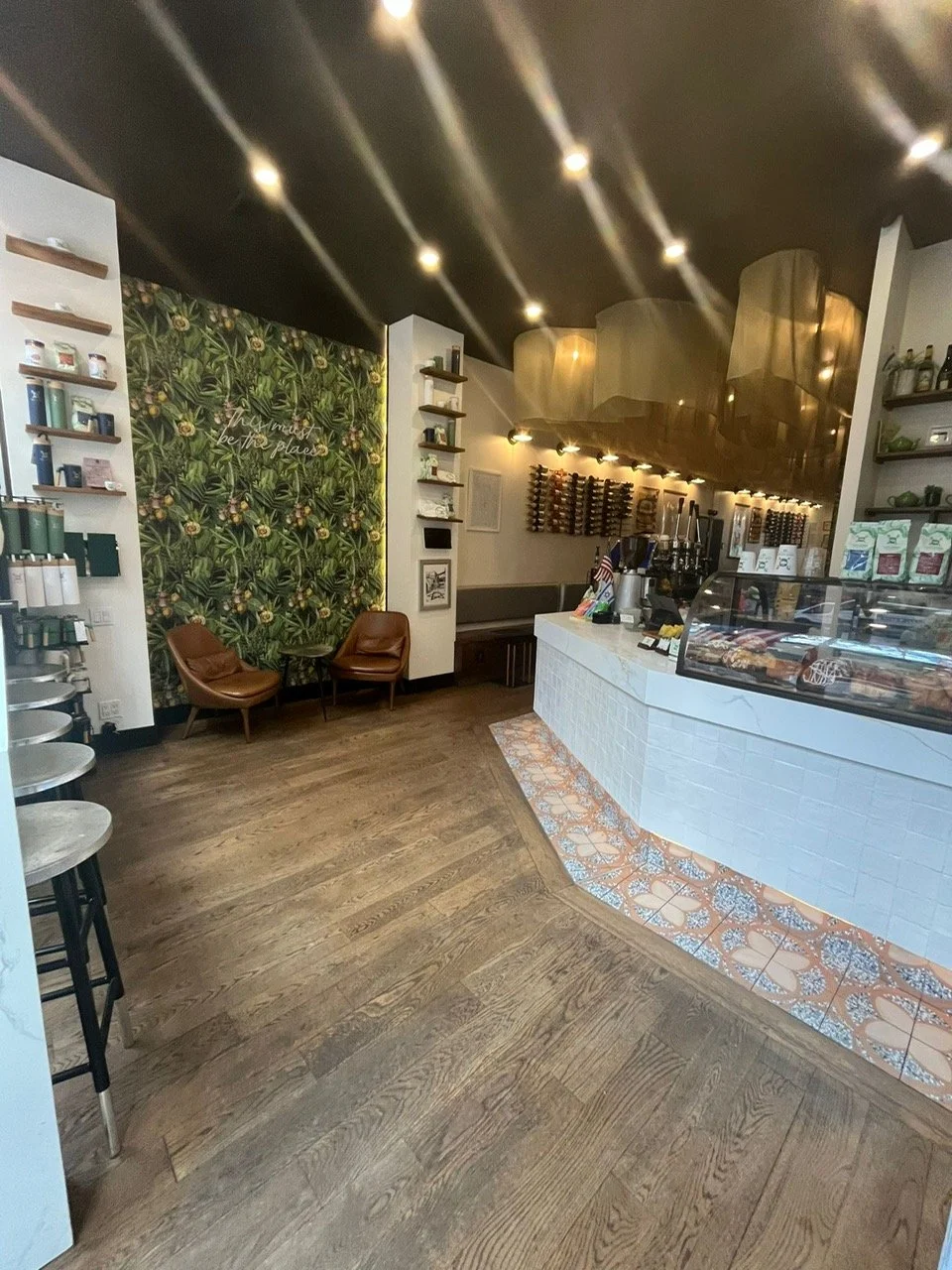 A modern, cozy cafe interior with a white counter, wooden flooring, green leafy wallpaper, and seating area with leather chairs.