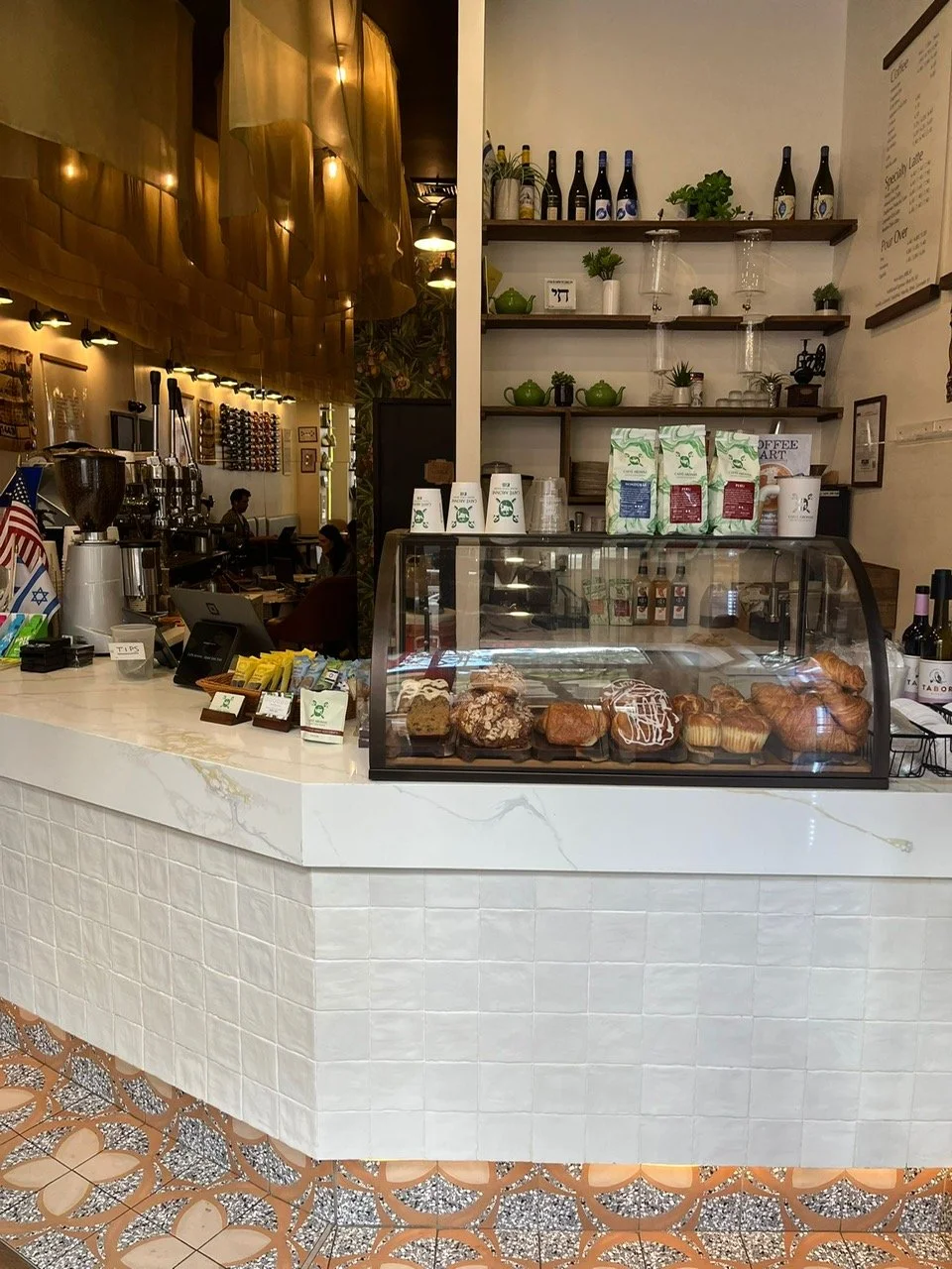 Counter of a coffee shop with a glass display case of assorted pastries, coffee bags, cups, and a shelf with wine bottles and decorative plants.