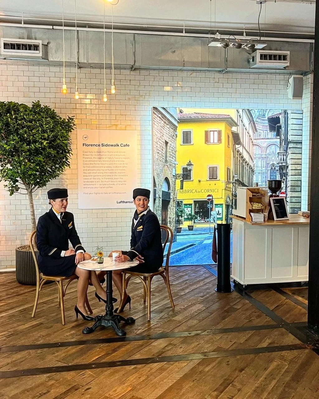 Interior of a cafe area with two flight attendant mannequins seated at a round table, a white brick wall with a sign, a potted tree, and a large window showing a street scene in Florence, Italy.