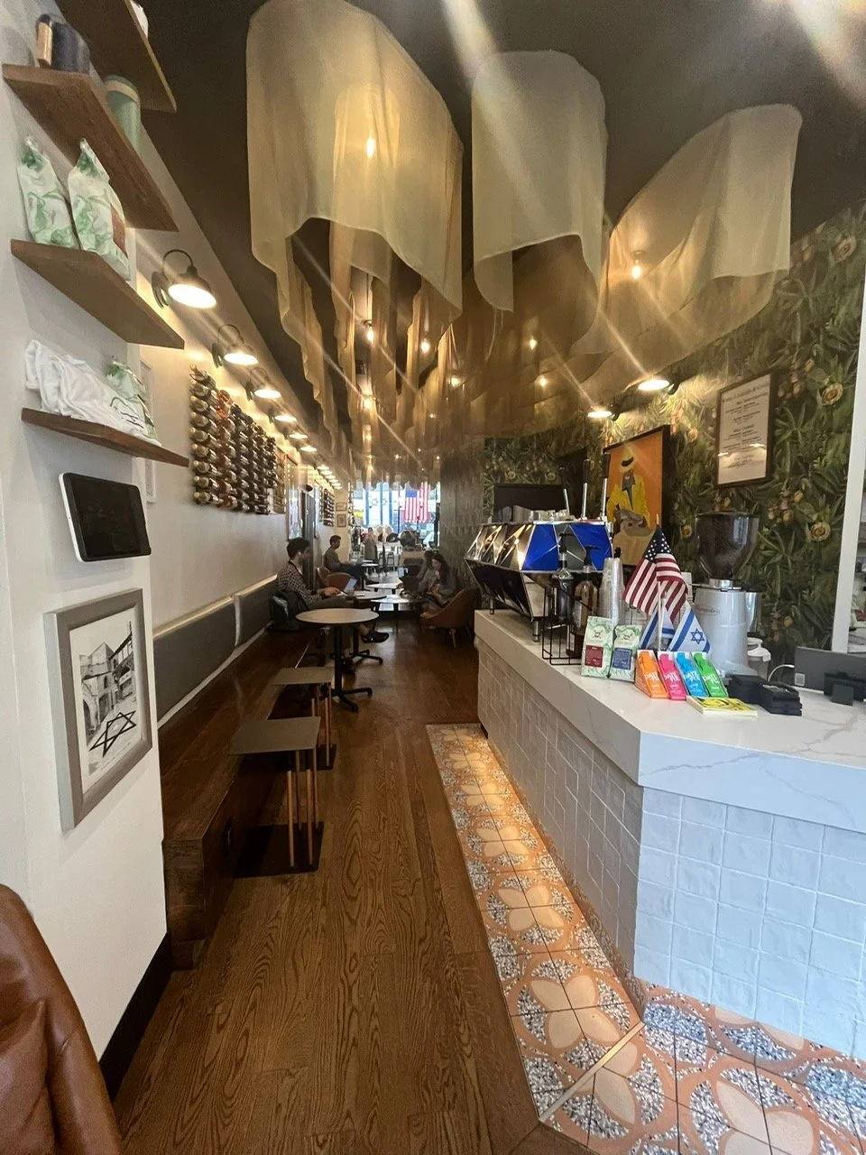 Interior of a cozy coffee shop with a white counter, Starbucks espresso machine, small American flags, colorful snack packages, and wall decorations. Customers sit at tables along a wall with wood flooring and decorative tiles near the counter.