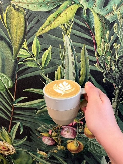 Hand holding a coffee cup with latte art in front of a tropical jungle mural with green leaves and plants.