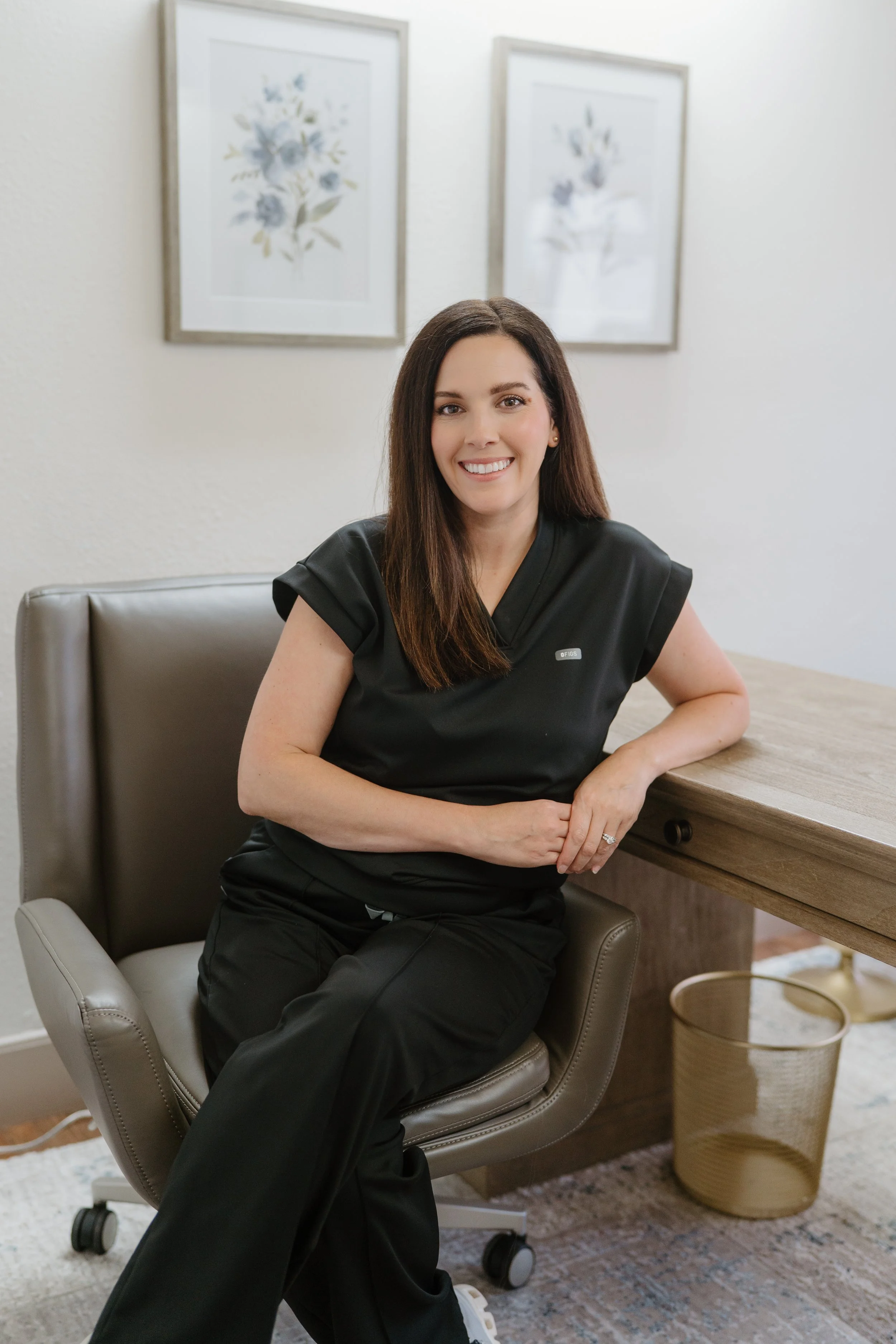 A woman with long brown hair, dressed in black scrubs, sitting in an office chair, smiling at the camera, with framed floral artwork on the wall behind her.