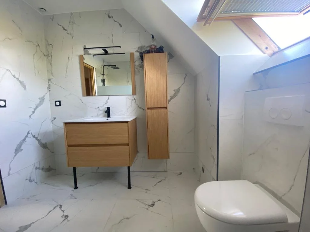 Salle de bain moderne avec murs et sol en marbre blanc à veines grises, lavabo en bois avec miroir au-dessus, cabine de douche, toilettes suspendues et un raccordement électrique au mur.