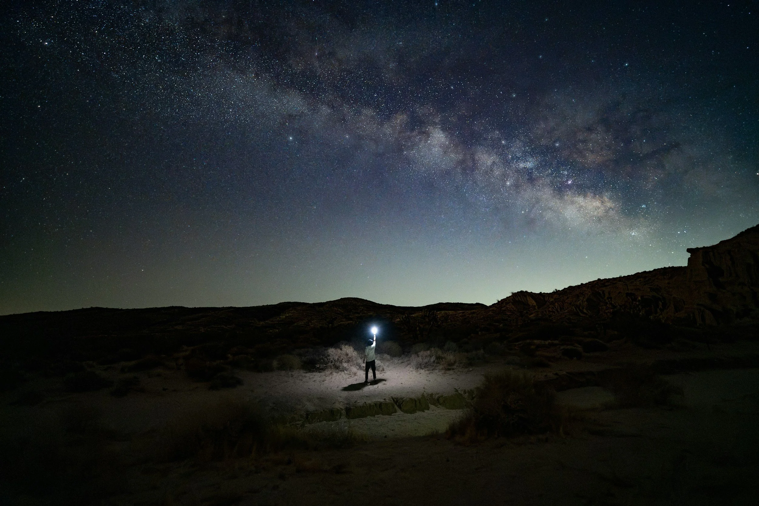Person standing in a desert under a starry night sky, holding a flashlight directed upwards.