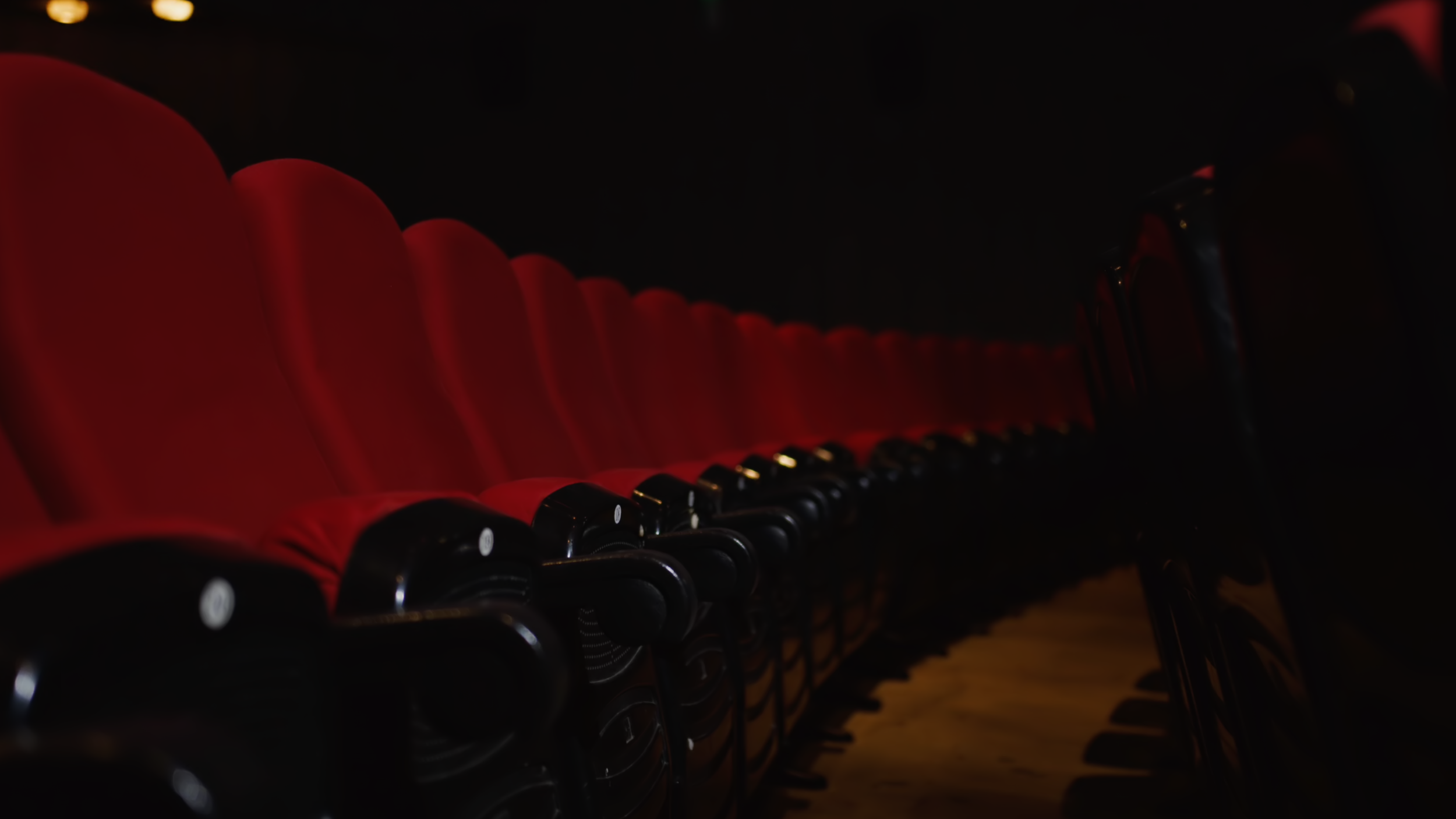Two young men sitting at a table in a cafe or restaurant, one holding a coffee cup and the other holding a drink with a straw, with a teal wall and menu in the background.