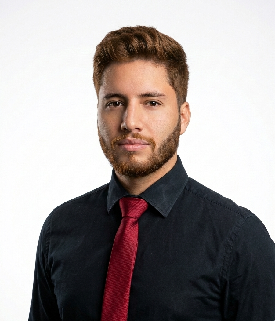A young man with light brown hair and a beard wearing a black dress shirt and a red tie, standing against a white background.