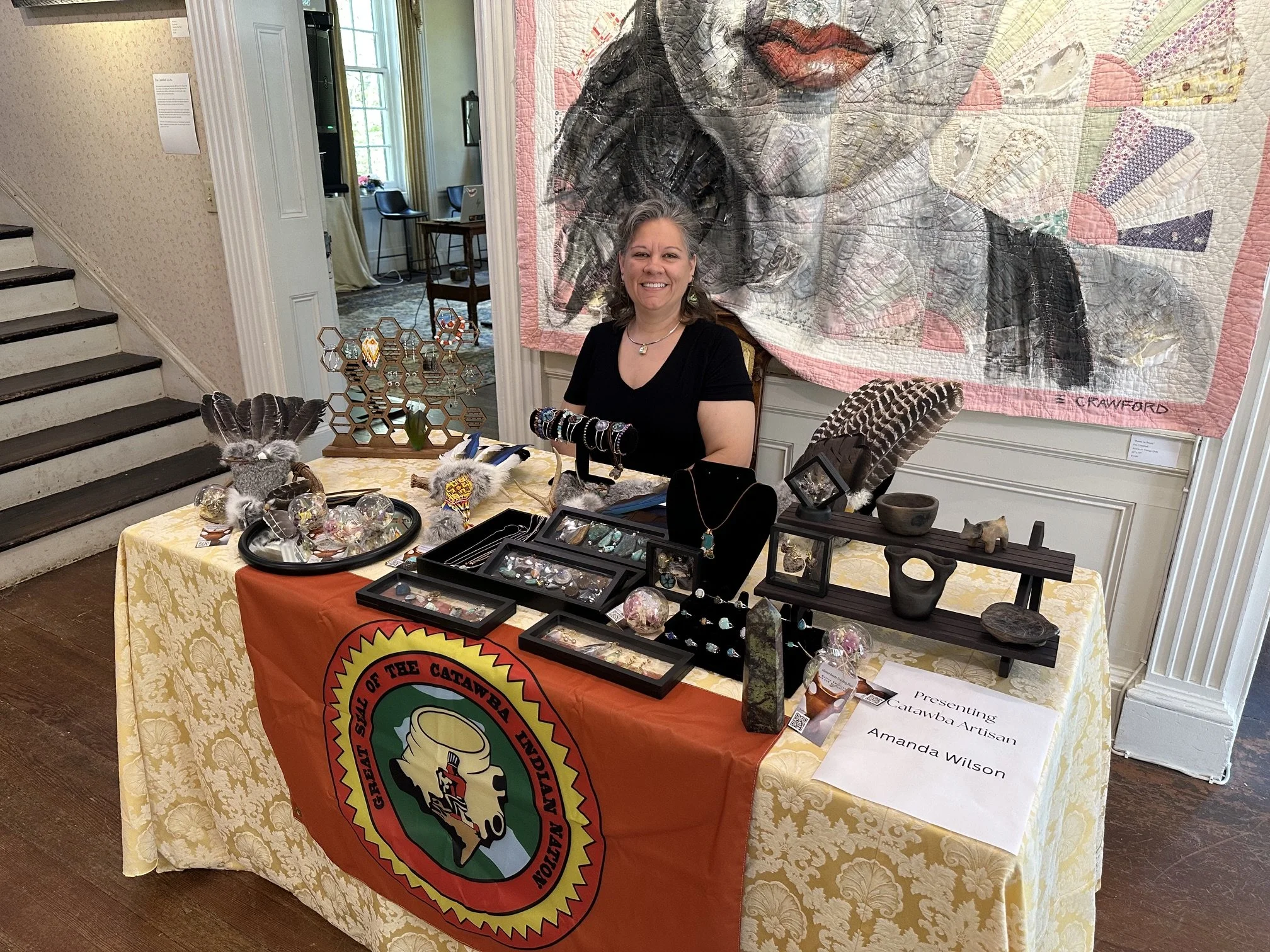 A woman sitting behind a display table at an indoor event, with jewelry and decorative objects. Behind her is a large quilted artwork of a woman's face. The table has an orange cloth with a logo, and the woman is smiling, wearing a black shirt and jewelry. There are stairs on the left and a room with a window and chairs in the background.
