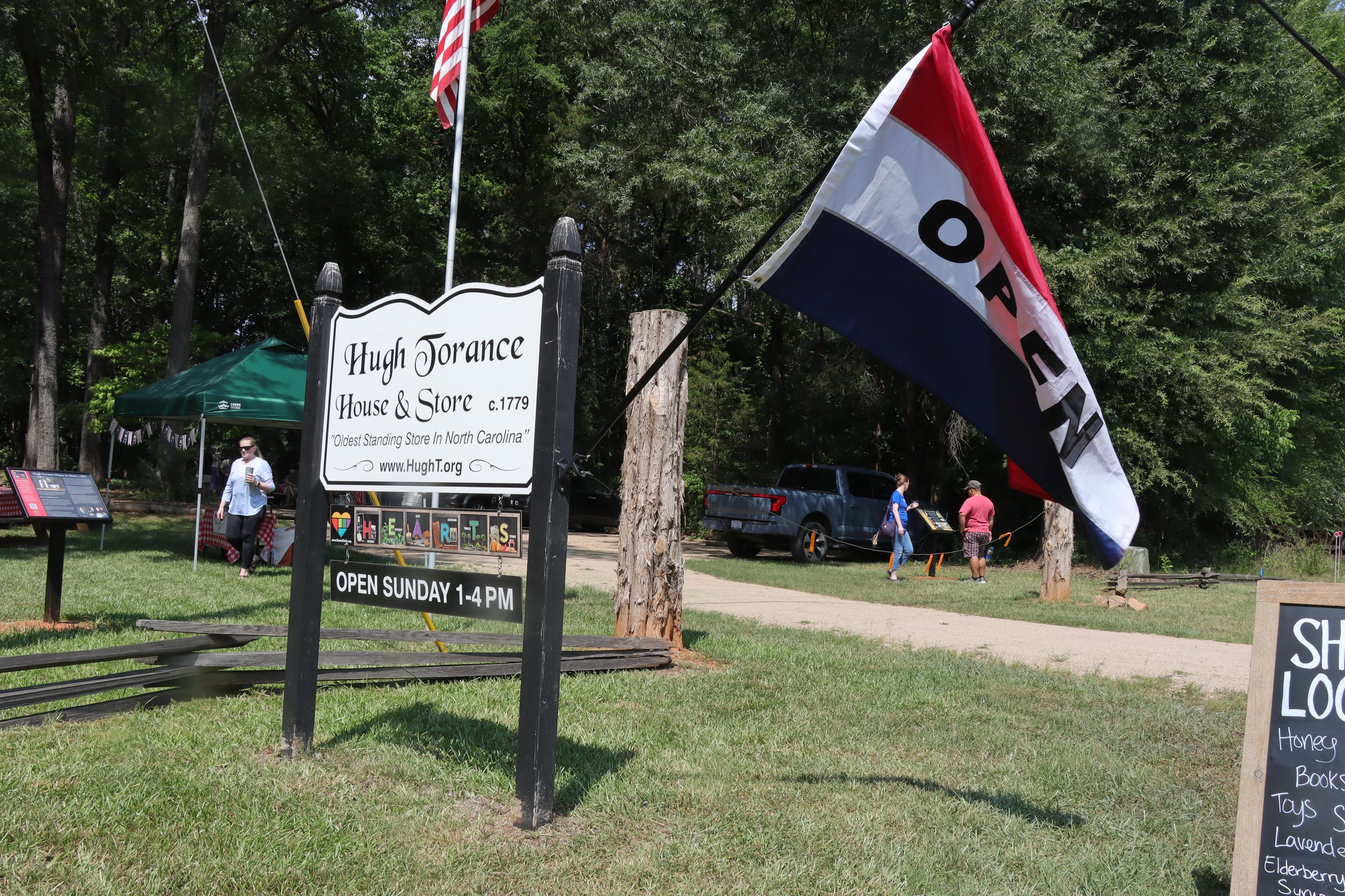 A park scene with people walking and a sign for the Hugh Torance House and Store, which is the oldest standing store in North Carolina. There is a flag with colors red, white, and blue, and a green canopy tent. The area has lush green trees and a grassy lawn.