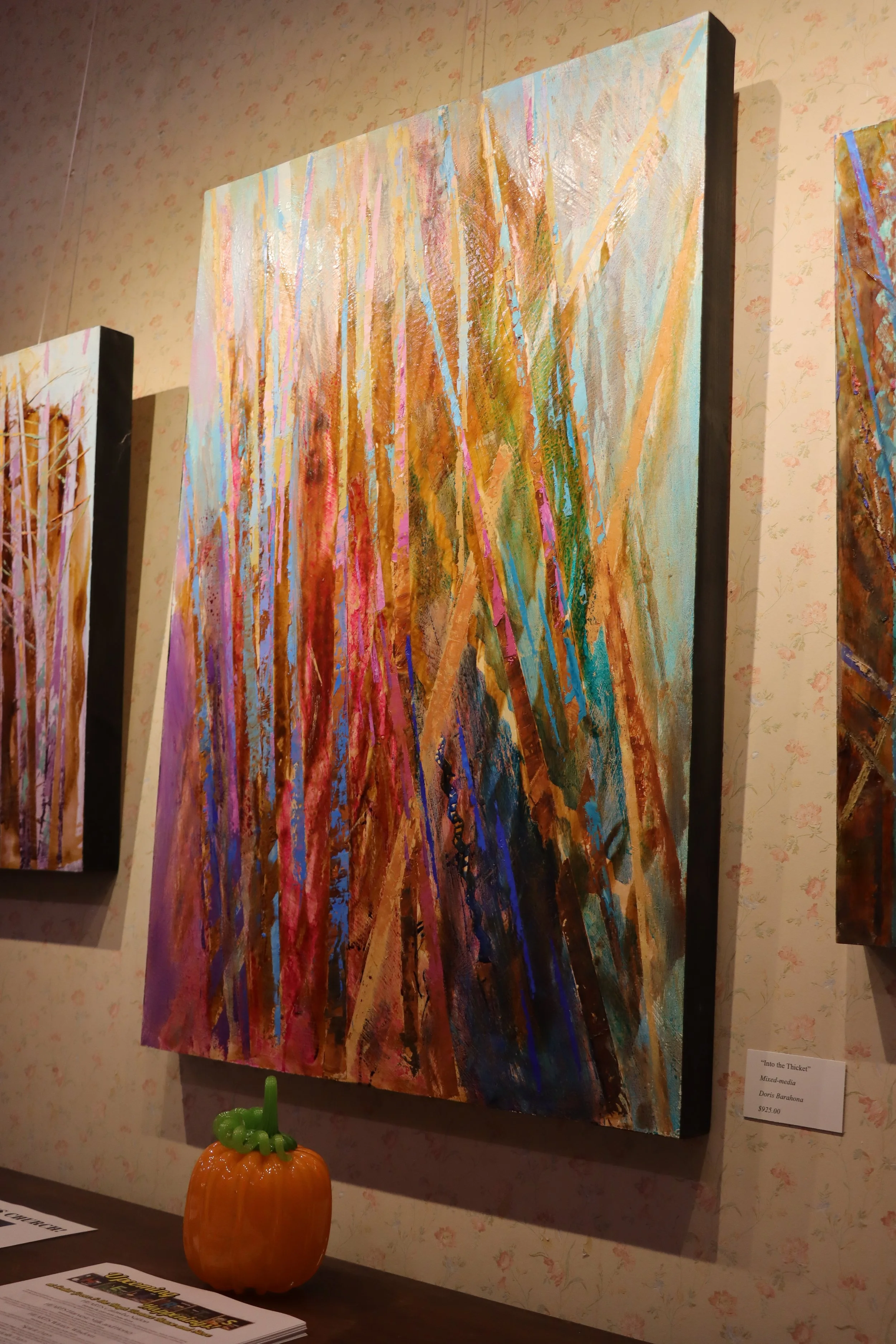 Colorful abstract mixed-media painting titled 'Into the Thicket' displayed on a gallery wall with a small pumpkin figurine on a table below and a printed description to the side.