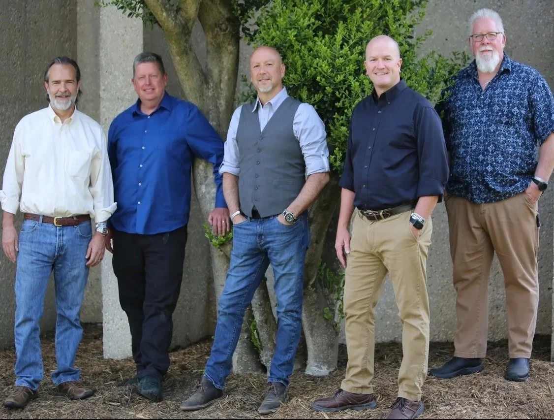 Five men standing outdoors in front of a tree and a concrete wall, smiling for a group photo.