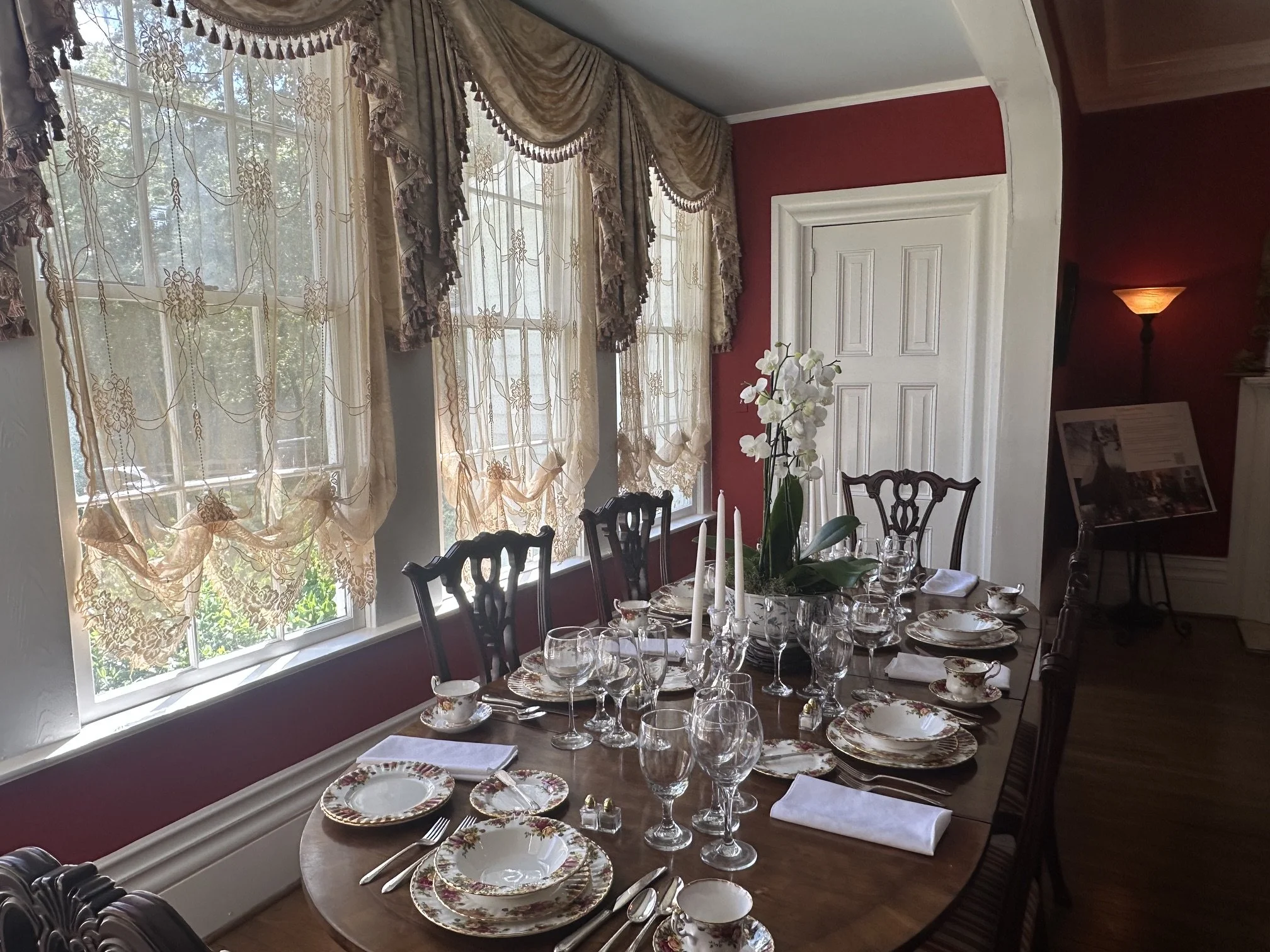 A dining room with a long wooden table set for a formal meal, featuring floral china, glassware, silverware, and white cloth napkins. The room has large windows with ornate lace curtains and draped valances, a white door, a red wall, and a table centerpiece with white orchids and tall candles. A lamp is lit in the background.
