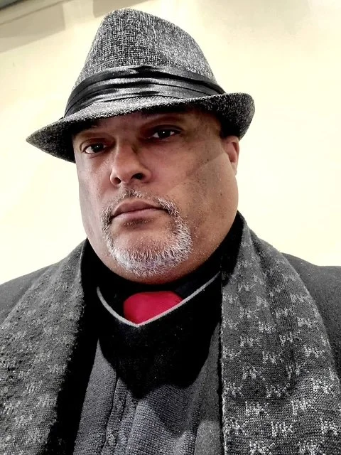 A man wearing a gray fedora hat with a black band, a black scarf with gray patterns, a black coat, and a black shirt with a red collar, looking serious.