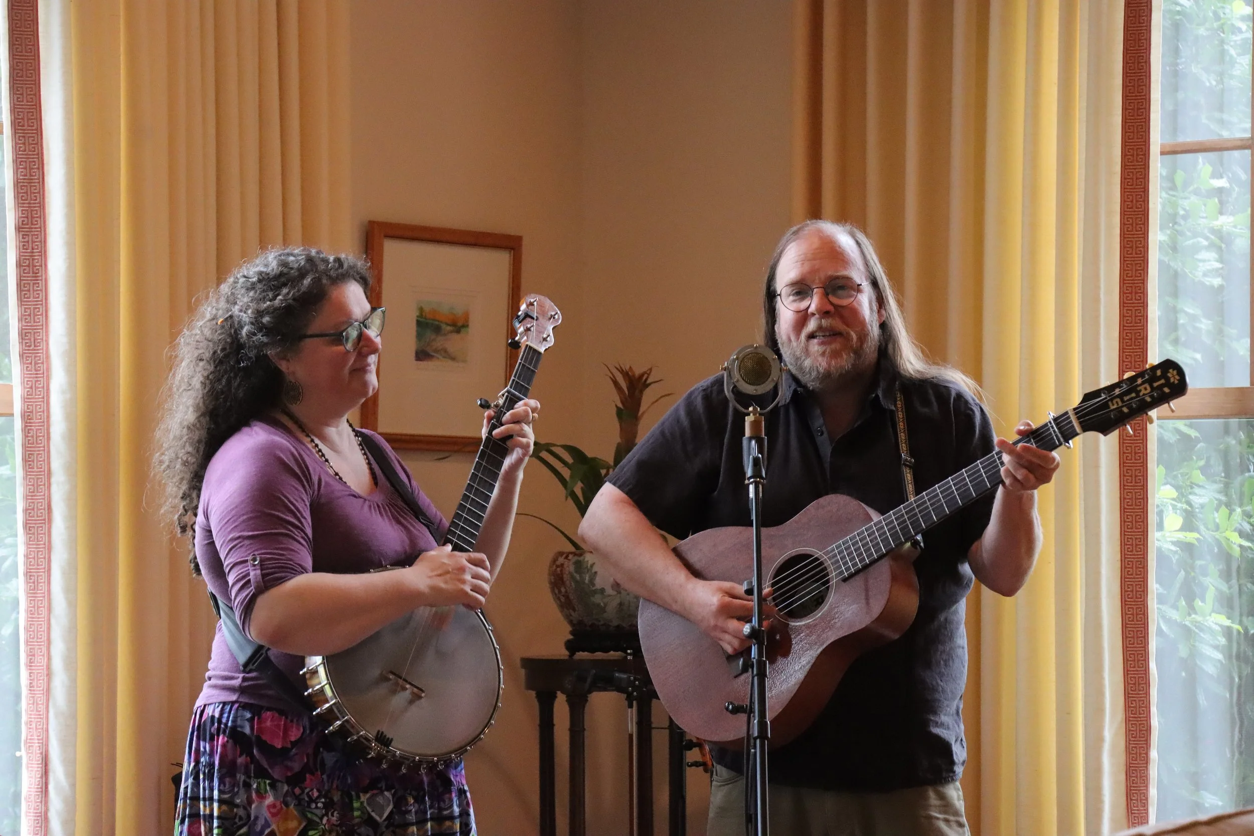 A woman and a man playing guitars and singing into a microphone in a room with yellow curtains and framed pictures on the wall.
