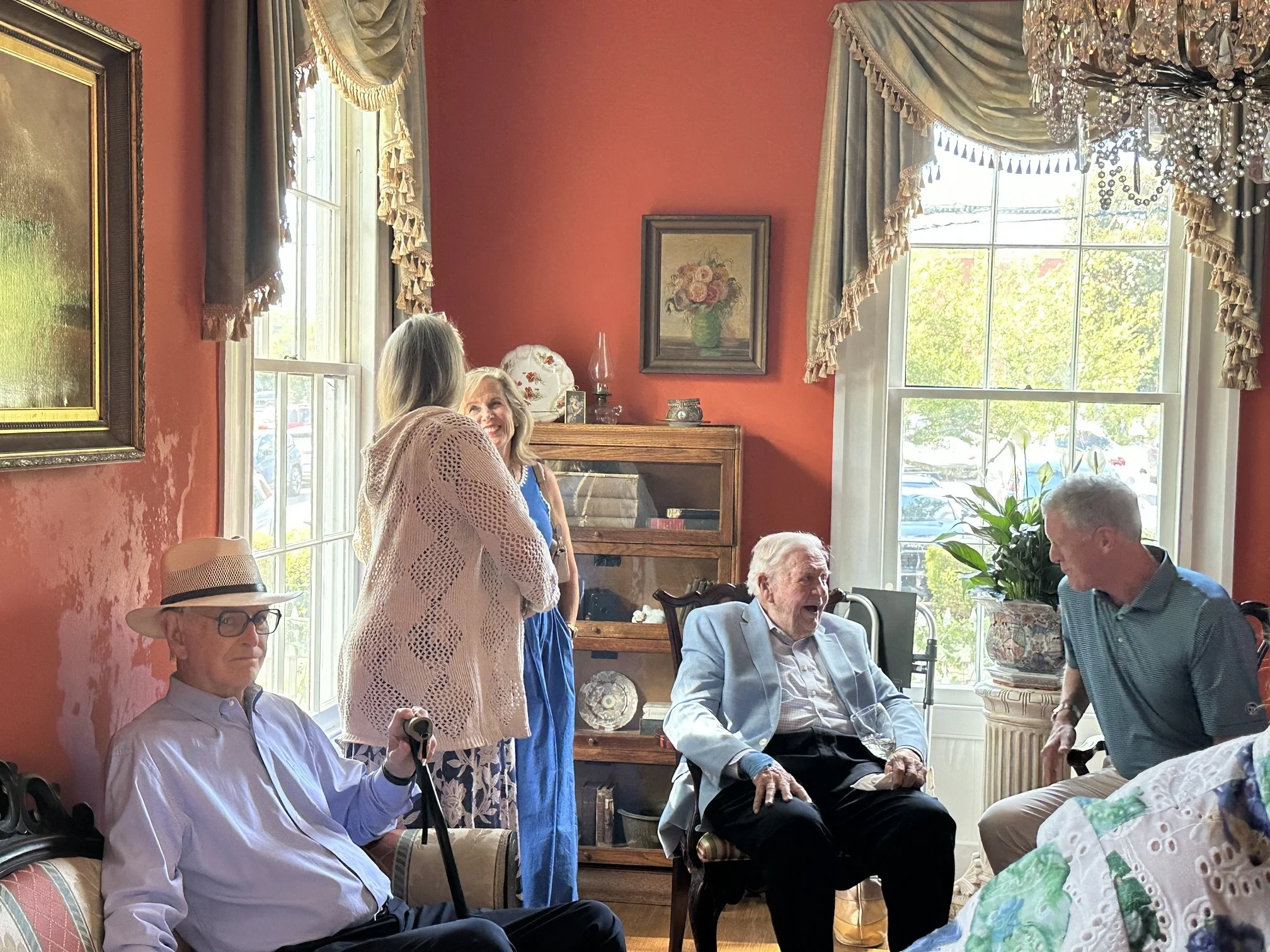 A group of elderly people and middle-aged adults gathered in a warmly lit living room with large windows, red walls, floral curtains, and framed artwork. Some are sitting on chairs, engaging in conversation, while others are standing and talking. The room is decorated with a waterfall of crystal beads and various decorative objects.