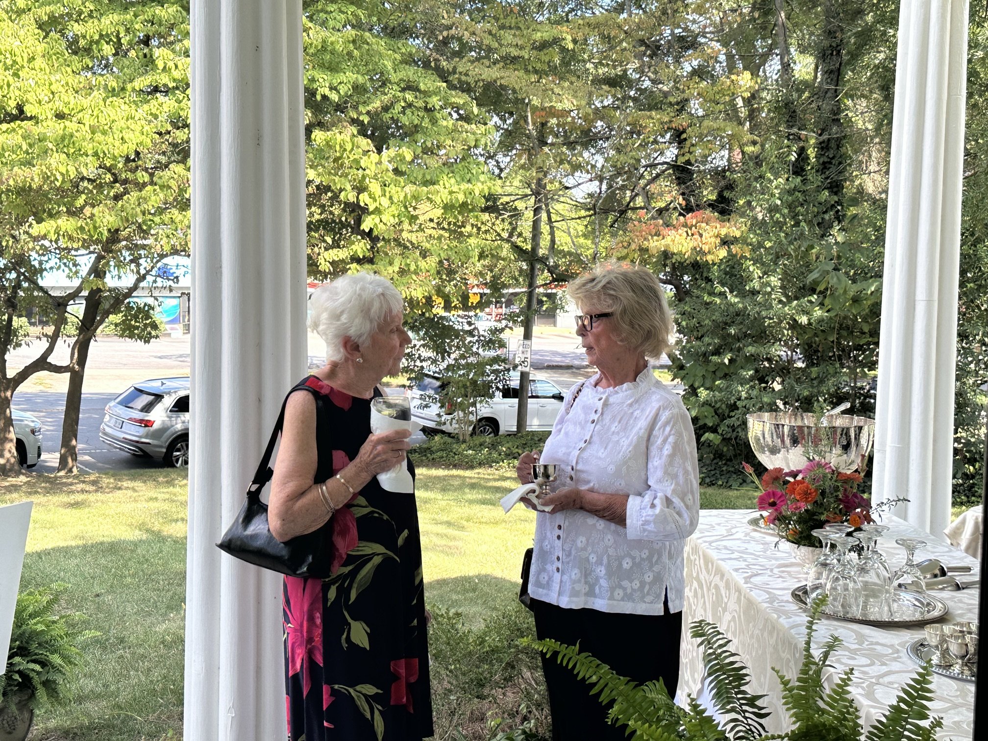 Two elderly women conversing at an outdoor event, one holding a glass of white wine and the other holding a silver cup, with a table of floral arrangements and silverware nearby and trees in the background.