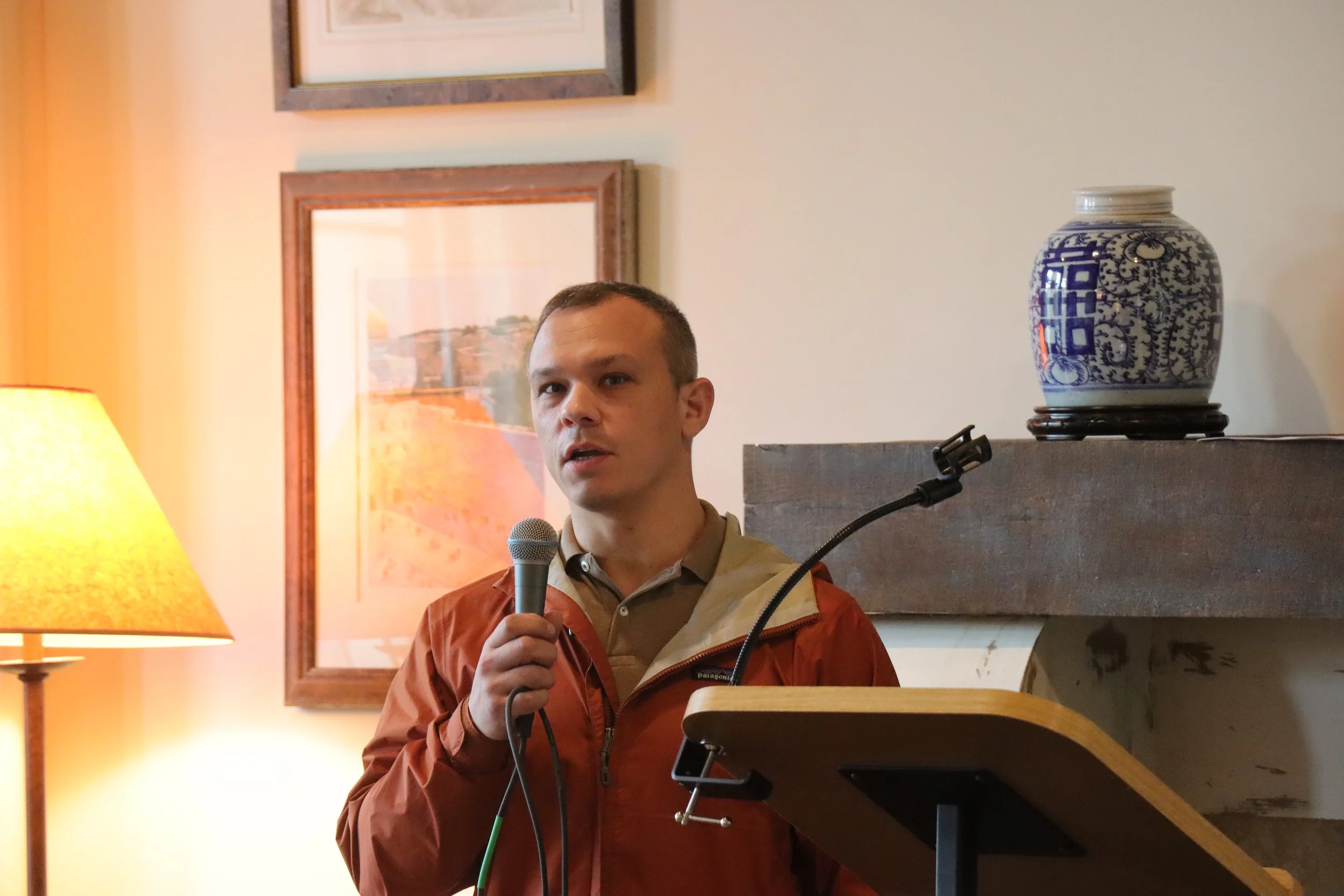 A man holding a microphone and speaking, standing indoors near a lectern with a blue and white decorated vase on a shelf behind him. There are framed paintings on the wall, a table lamp, and framed pictures in the background.