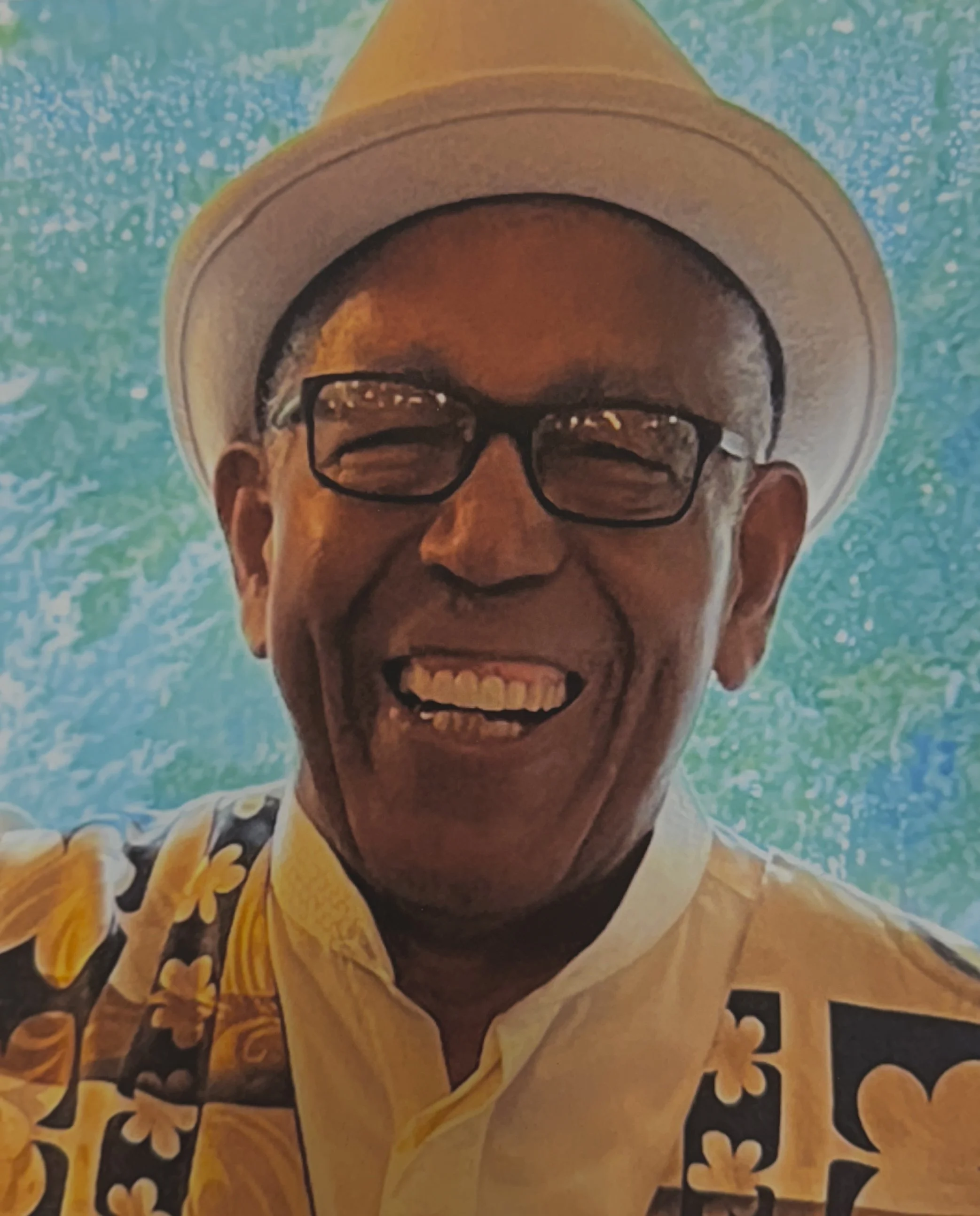 A smiling elderly man wearing glasses, a white hat, and a yellow Hawaiian shirt with black and yellow flower patterns, standing in front of a body of water with turquoise waves.