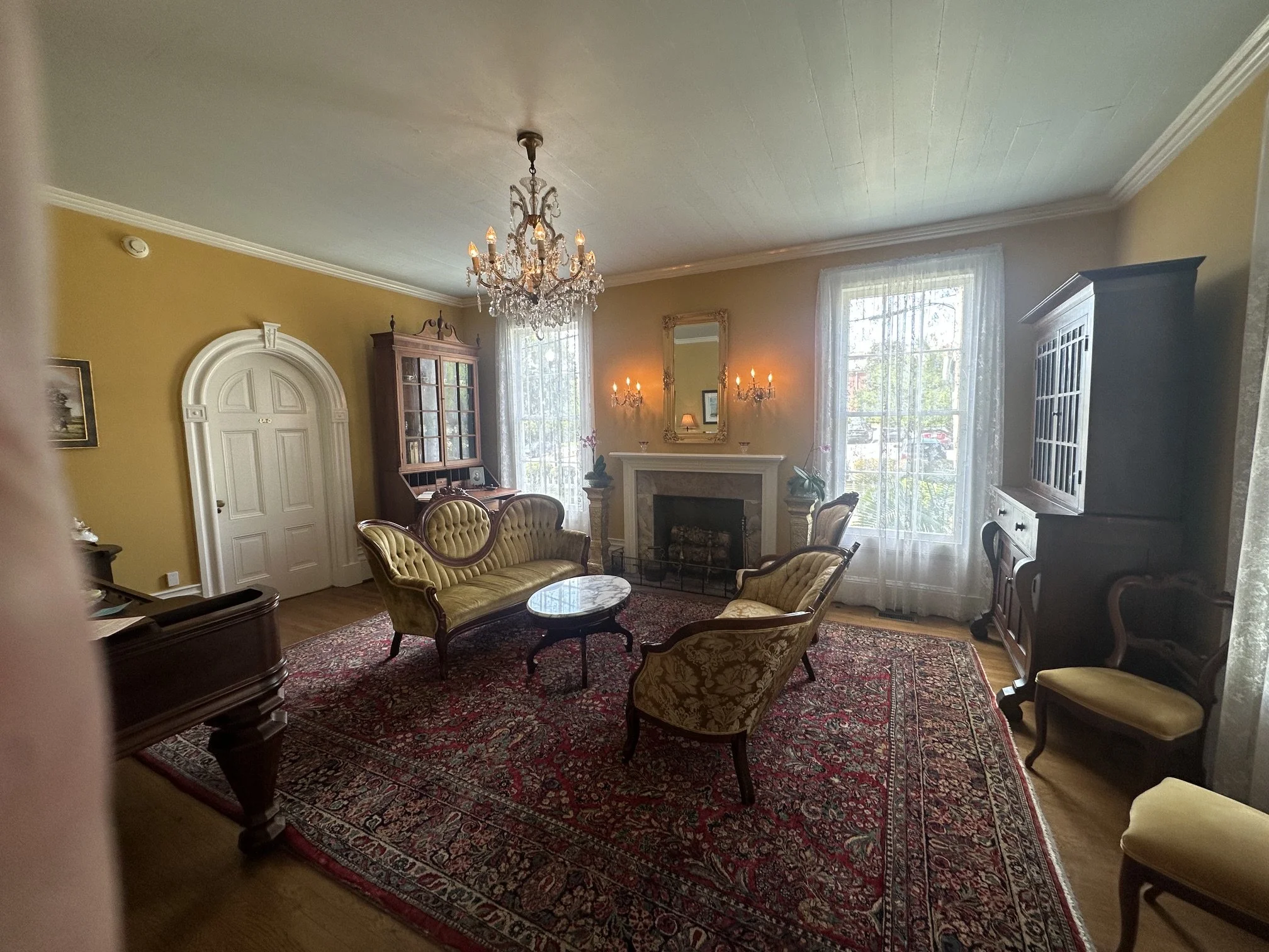 Living room with vintage furniture, chandelier, fireplace, large windows with sheer curtains, yellow walls, and ornate mirror above the fireplace.