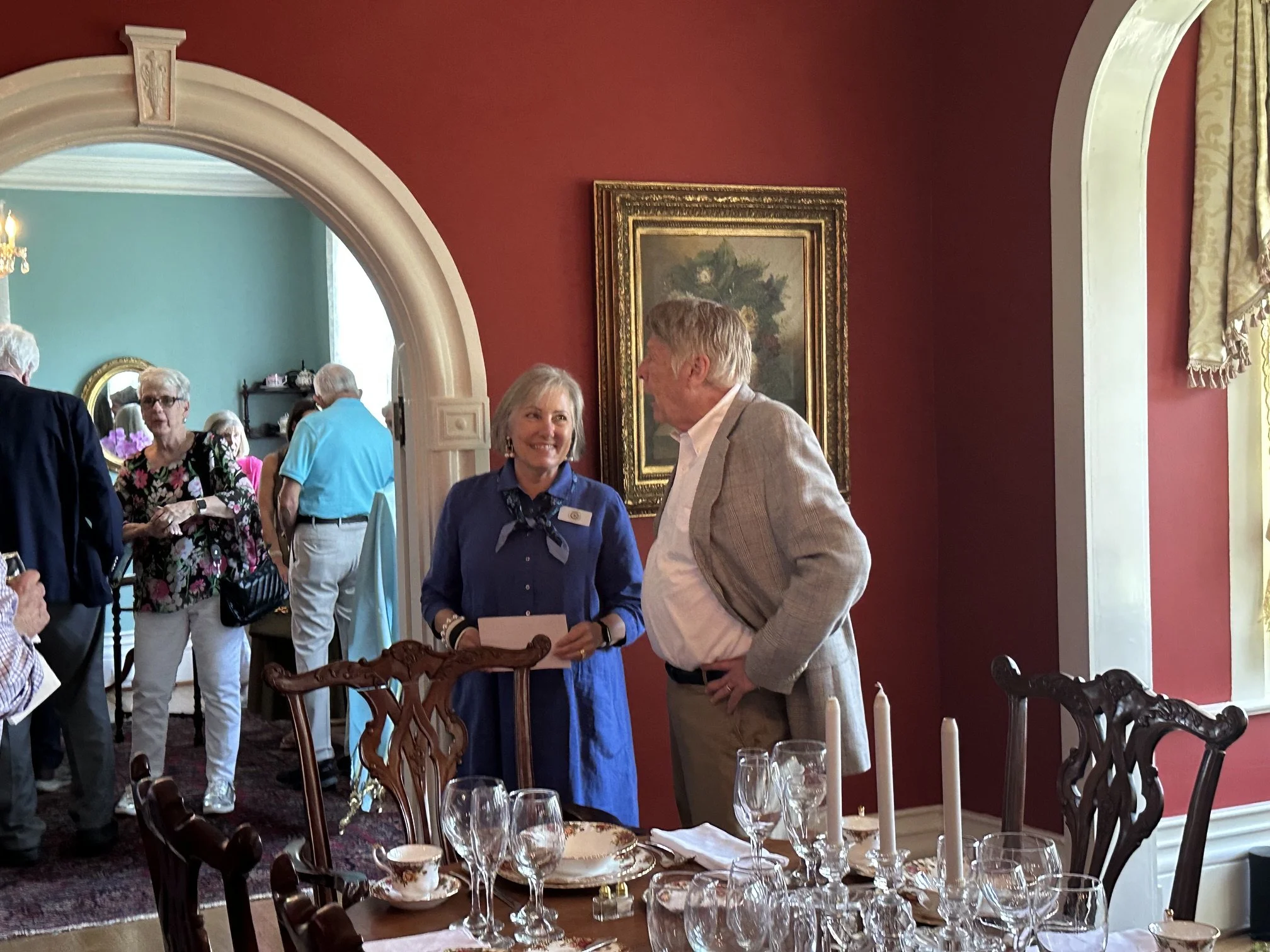 An indoor gathering with people engaged in conversation, in a room with red walls and a decorated dining table set with wine glasses, plates, and candlesticks. Visible artwork on the wall and a separate room with blue walls and floral decor.
