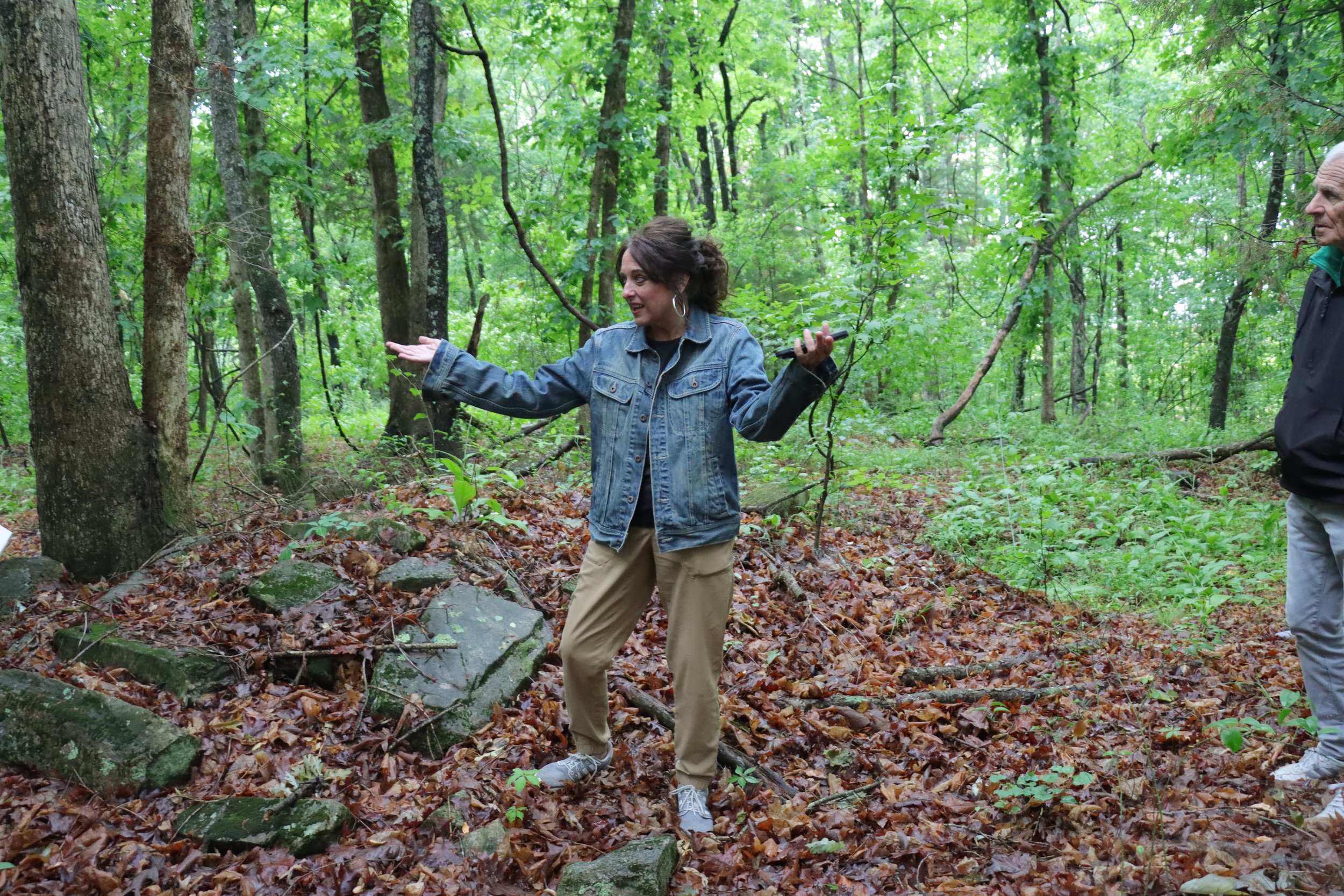 Two women and a man in a dense green forest. The woman in the middle is gesturing with her hands, and the man on the right is looking at her. The forest floor is covered with fallen leaves, rocks, and vegetation.