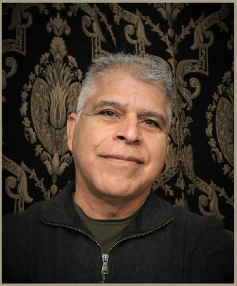 A middle-aged man with short gray hair, smiling, wearing a black zip-up jacket and a dark green shirt, standing in front of a ornate black and gold patterned background.