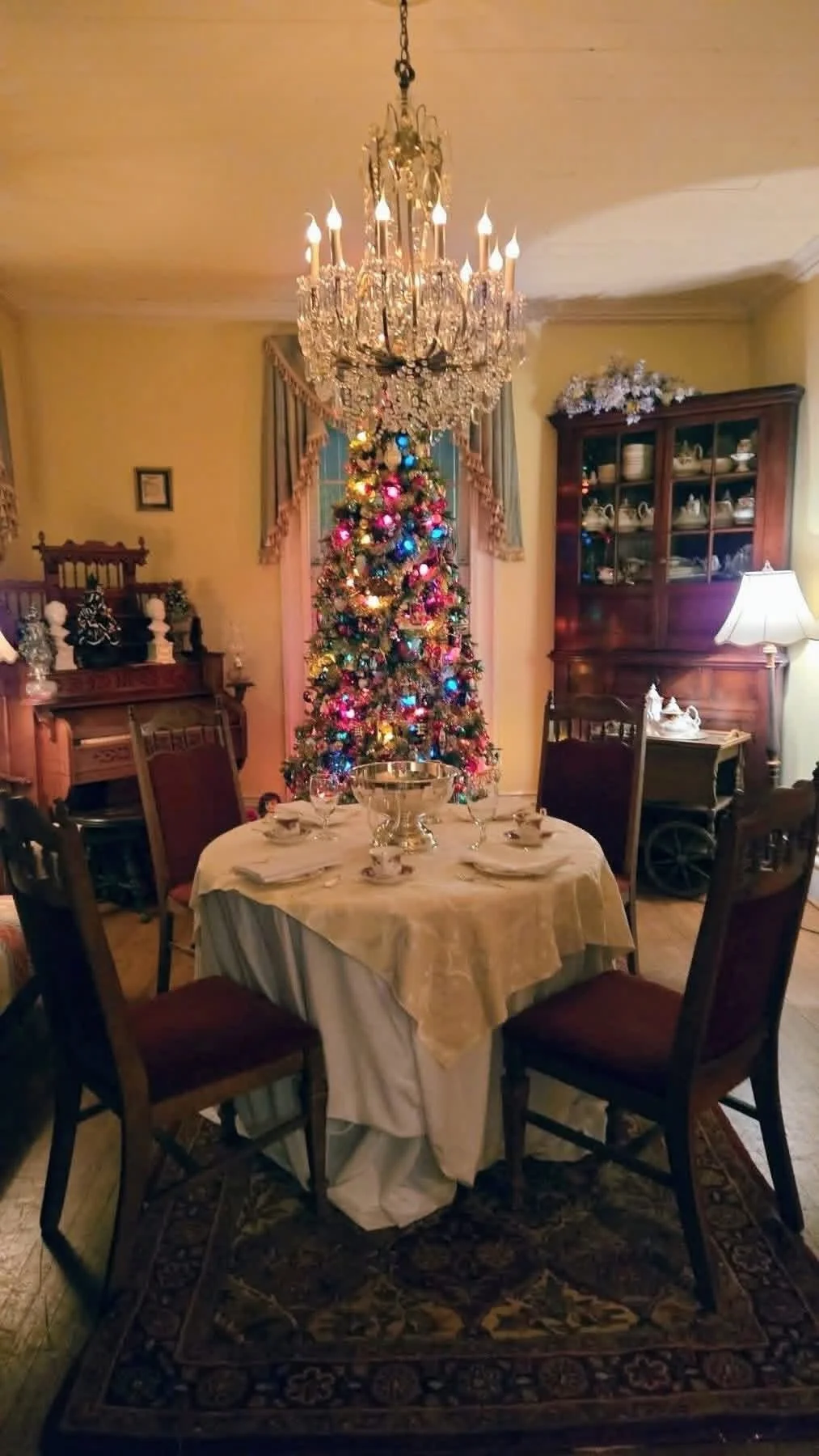 A decorated Christmas tree with colorful lights in a dining room, with a chandelier above, a table set with dishes, and antique furniture surrounding the room.