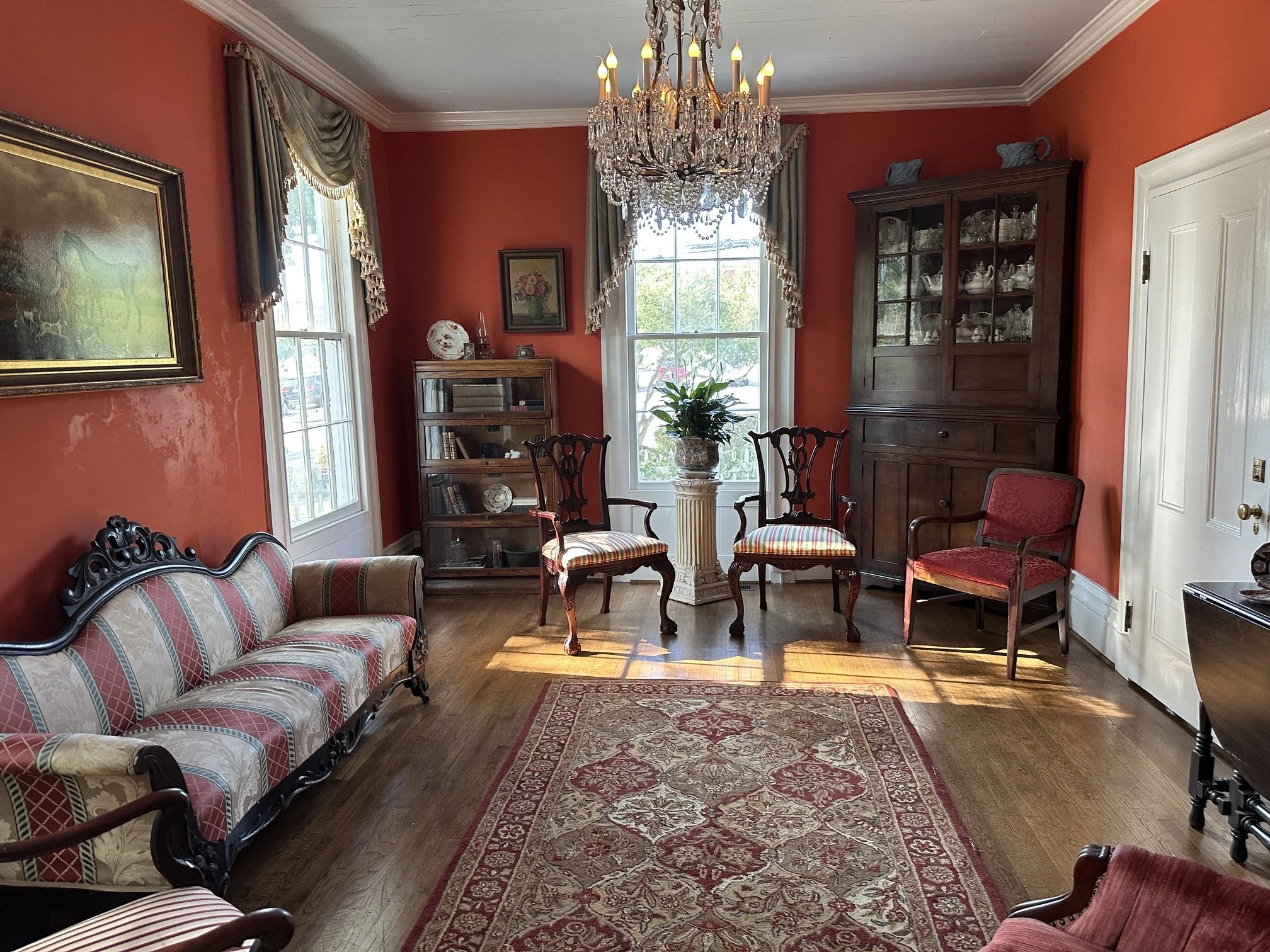 A formal living room with red walls, a chandelier, large windows with curtains, a wooden china cabinet, a small bookshelf, a plant in a decorative planter, a patterned area rug, and various antique-style chairs and a sofa.