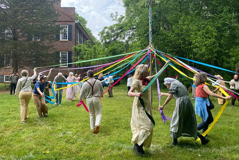 People dressed in vintage clothing participate in a maypole dance outdoors on a grassy lawn, holding colorful ribbons attached to a tall pole, surrounded by trees and a brick building in the background.