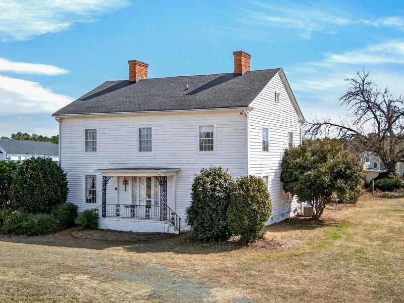 A white two-story house with a gray roof, surrounded by bushes and trees, under a blue sky.