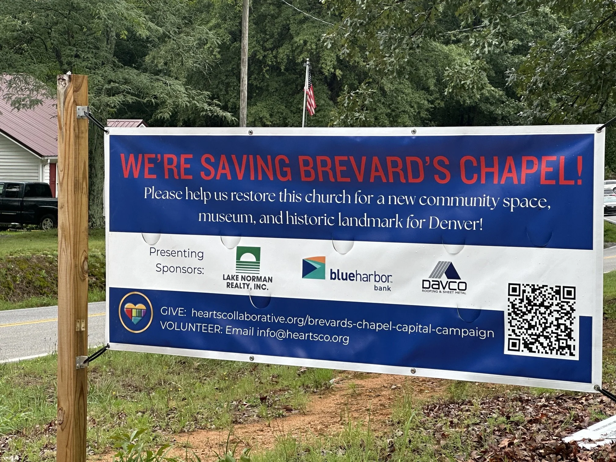 A large blue and white campaign sign with red text that says, 'We're saving Brevard's Chapel!' The sign includes a message requesting help for restoring a church for a community space, museum, and historic landmark in Denver. The sign displays logos of sponsors: Lake Norman Realty, Blue Harbor Bank, and Daco Roofing & Sheet Metal. It also has a QR code and contact information for donations and volunteer efforts. The sign is mounted on a wooden post, with trees, a house, and a flag in the background.