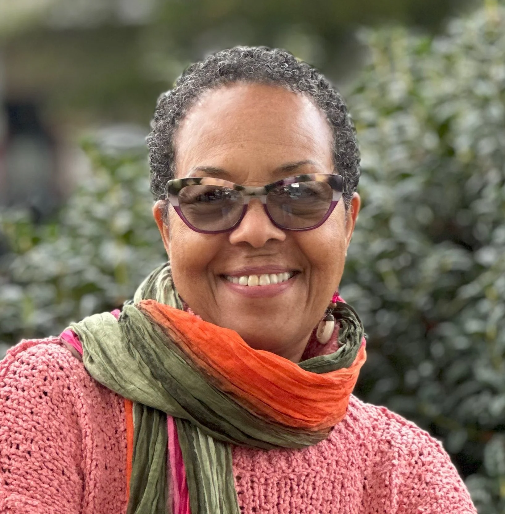 Smiling woman with short curly gray hair wearing sunglasses, a pink textured sweater, and a multicolored scarf outdoors with a blurred green background.