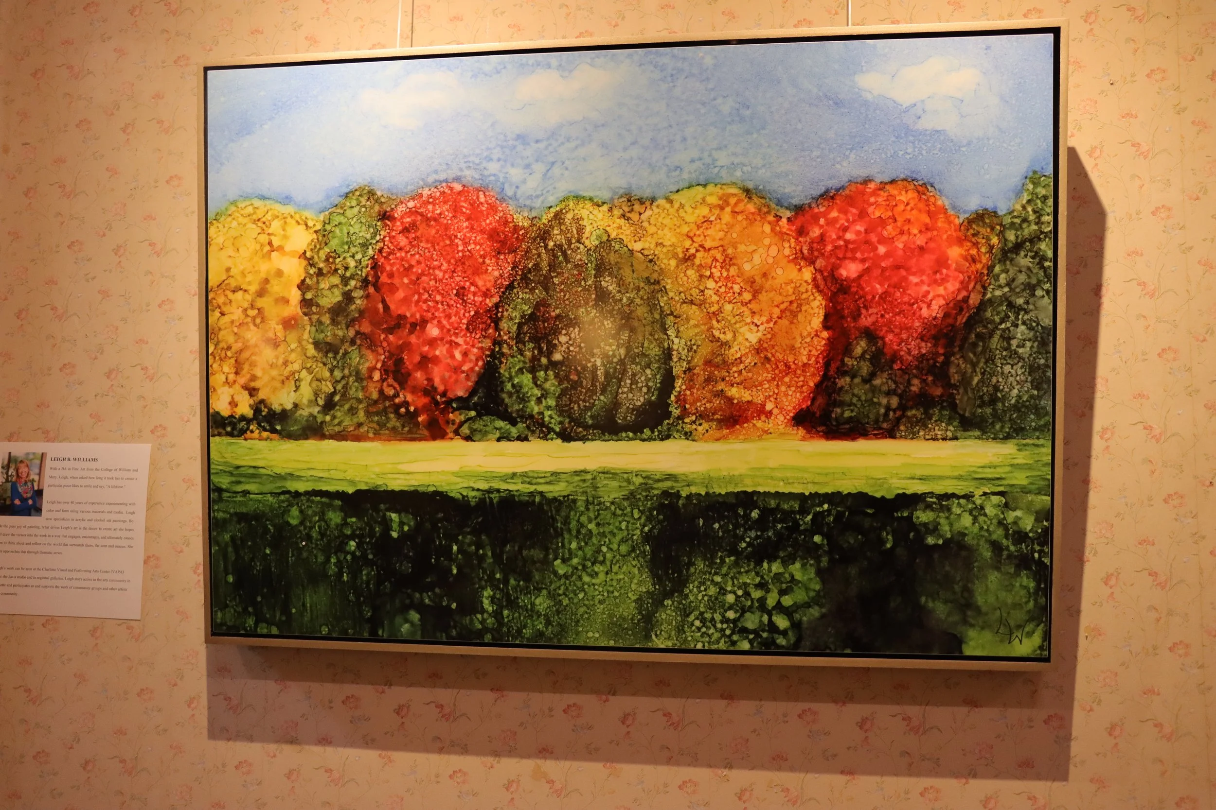 Colorful landscape painting featuring a line of multicolored autumn trees with red, yellow, orange, green, and dark foliage, set against a blue sky with a few clouds, and a green grassy foreground.