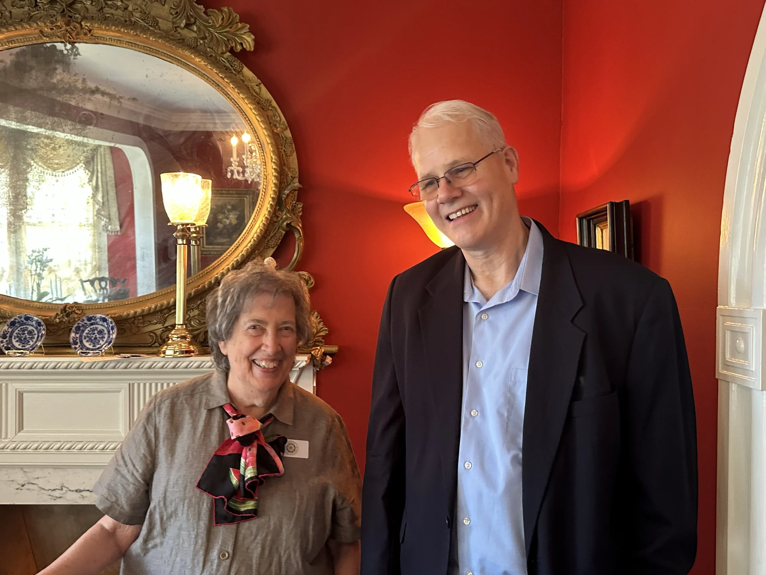 A man and a woman smiling indoors with a red wall, ornate mirror, and a decorative lamp behind them.