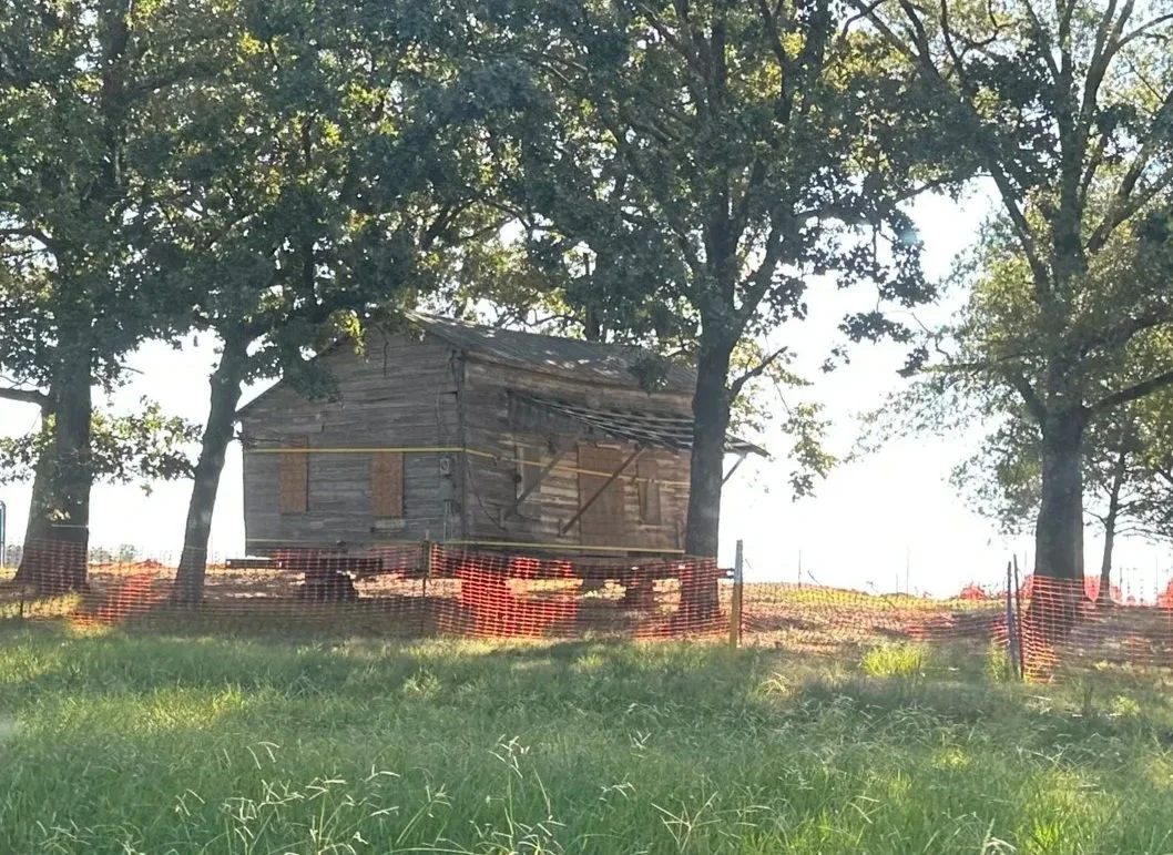 Old, weathered wooden barn on a grassy hill surrounded by trees with sunlight shining through the leaves.