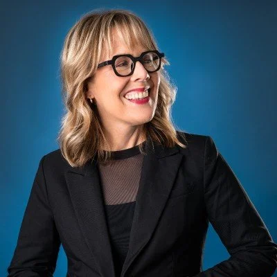 A woman with blonde hair wearing glasses and a black blazer, smiling against a blue background.