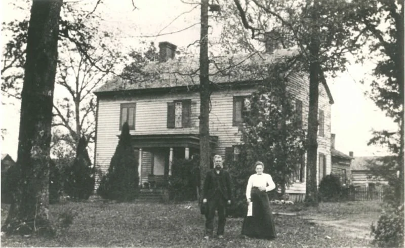 A white two-story house with black shutters and a front porch, surrounded by green bushes and trees, under a clear blue sky.