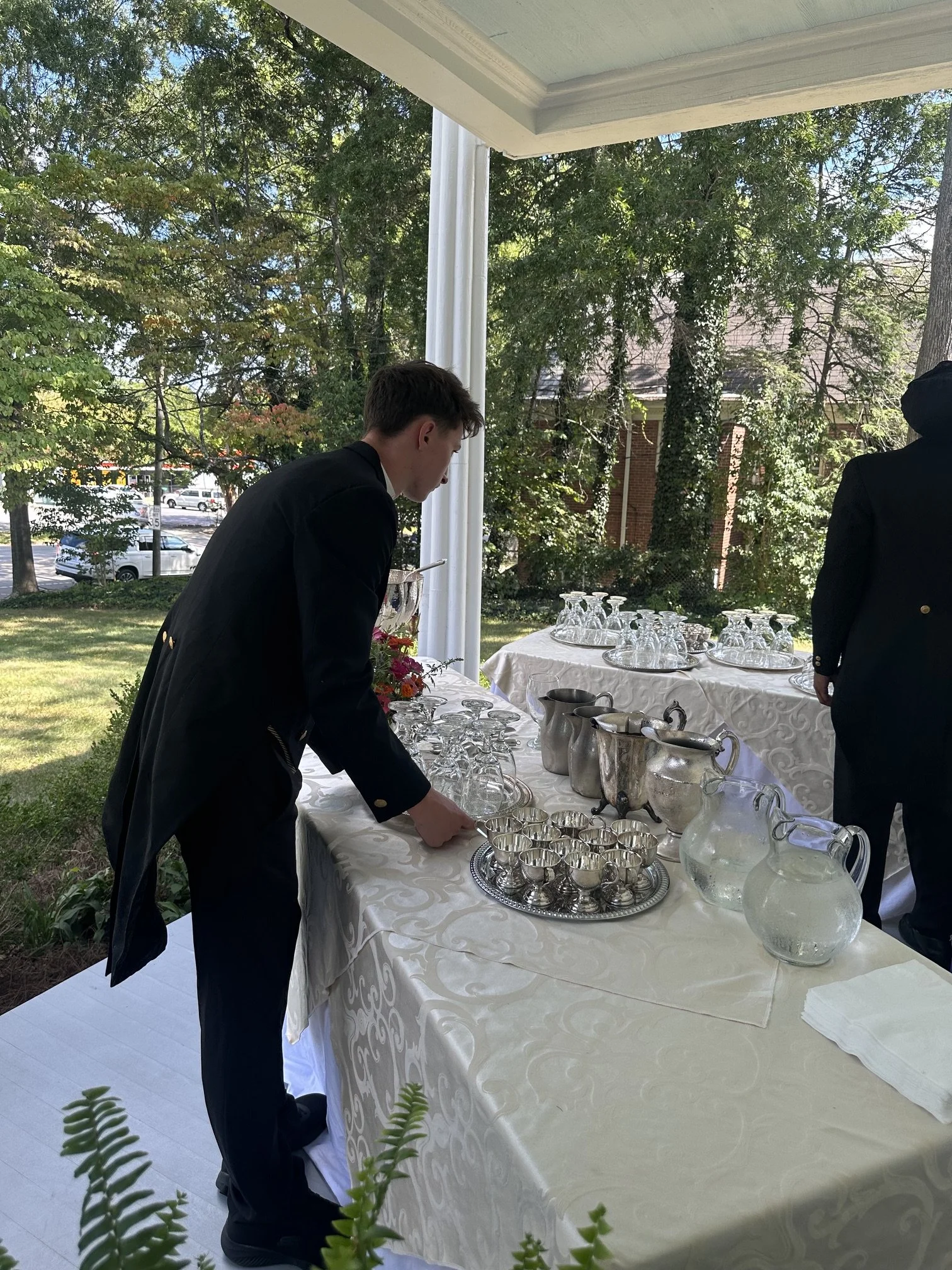 A young man in a black suit and white shirt is setting up glasses on a decorated table at an outdoor event under a white tent with trees and a building in the background.