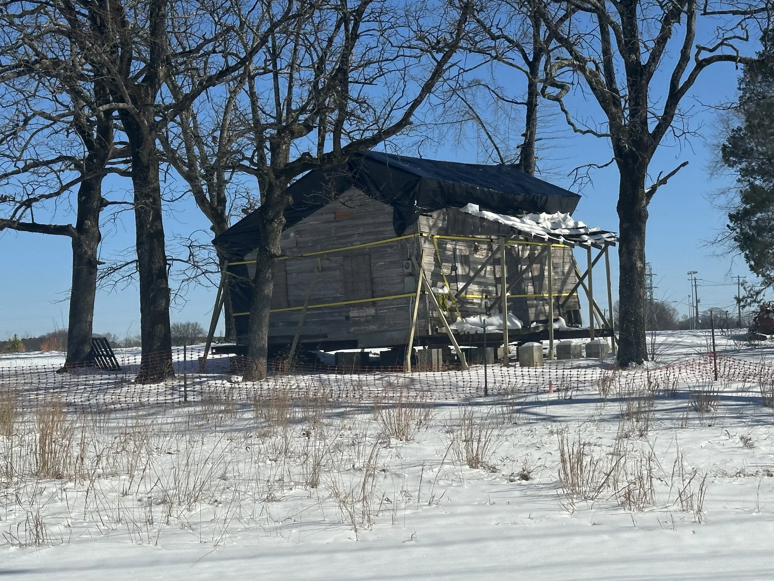 A small wooden house elevated on stilts surrounded by snow, with a black tarp covering part of the roof, set among leafless trees on a cold winter day.