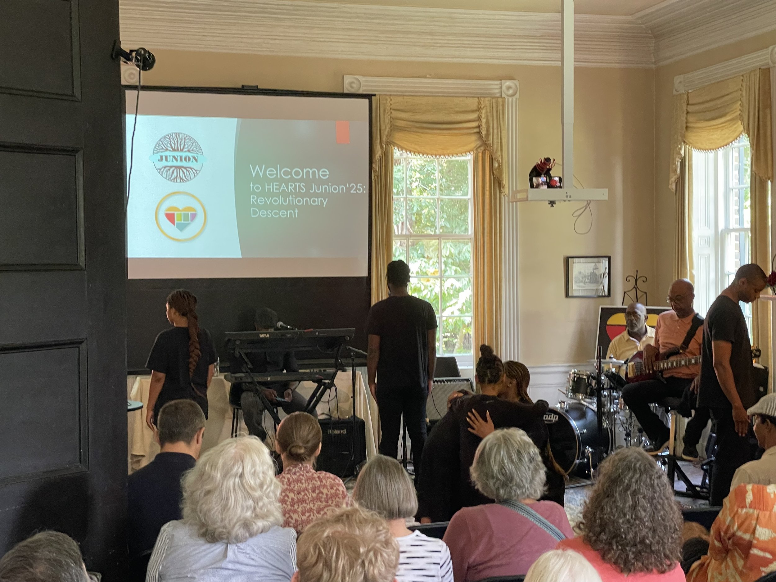 A large group of people seated in a room, facing a presentation screen. The screen displays the logo and text for 'HEARTS Junion 25: Revolutionary Descent'. The room has large windows with yellow curtains, and there is musical equipment and a projector mounted on the wall.