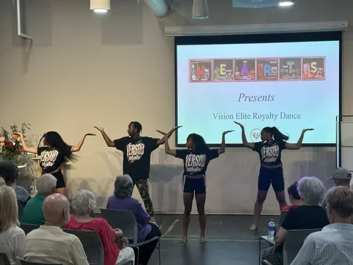 Five young dance performers in black shirts with white lettering, dancing on stage in front of an audience, with a large screen behind them displaying 'HELLO STUDENTS Presents Vision Elite Royalty Dance.'