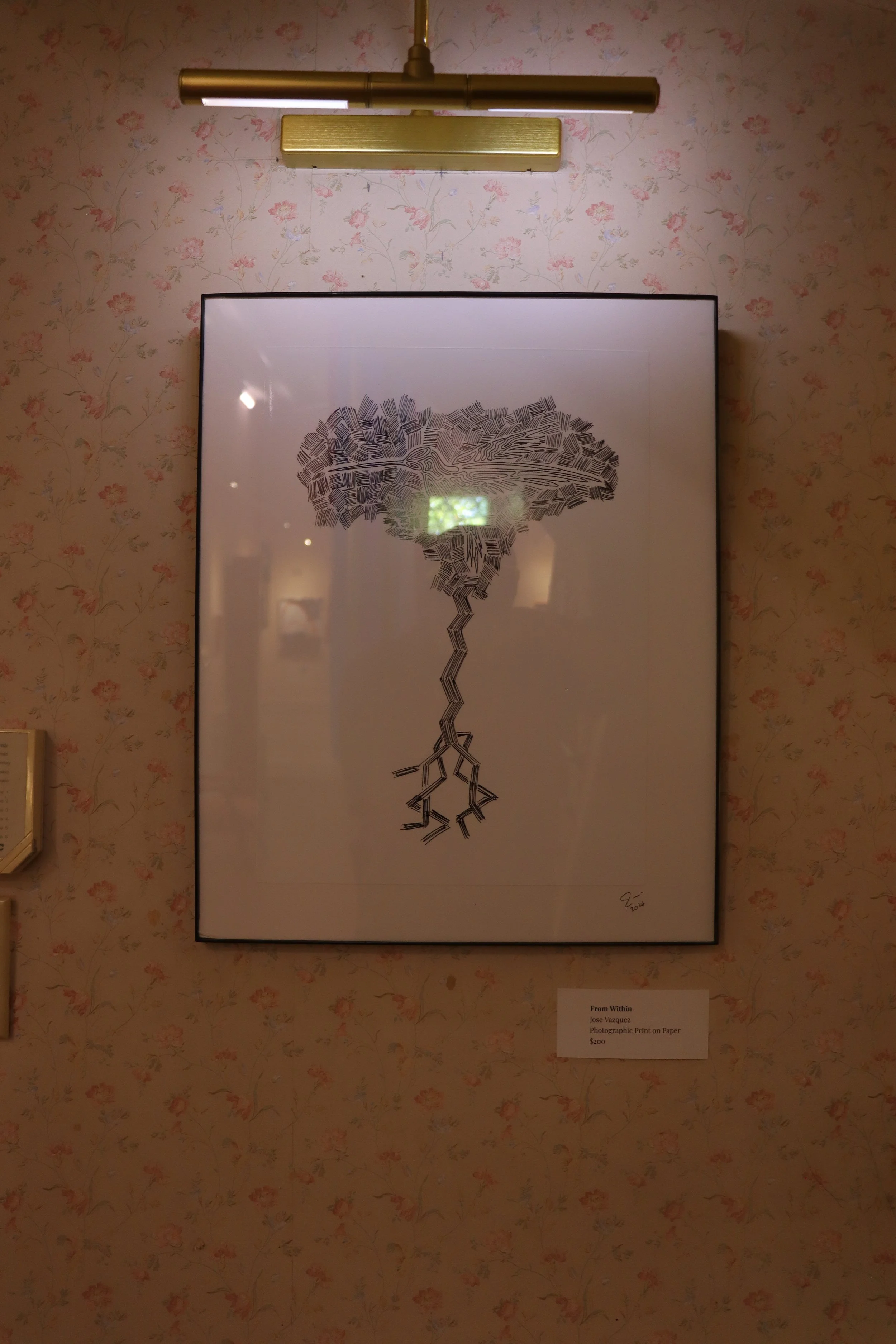 A framed artwork depicting a stylized human brain composed of black lines and patterns, with a stem-like extension at the bottom, hanging on a floral wallpapered wall under a metallic wall-mounted light fixture.