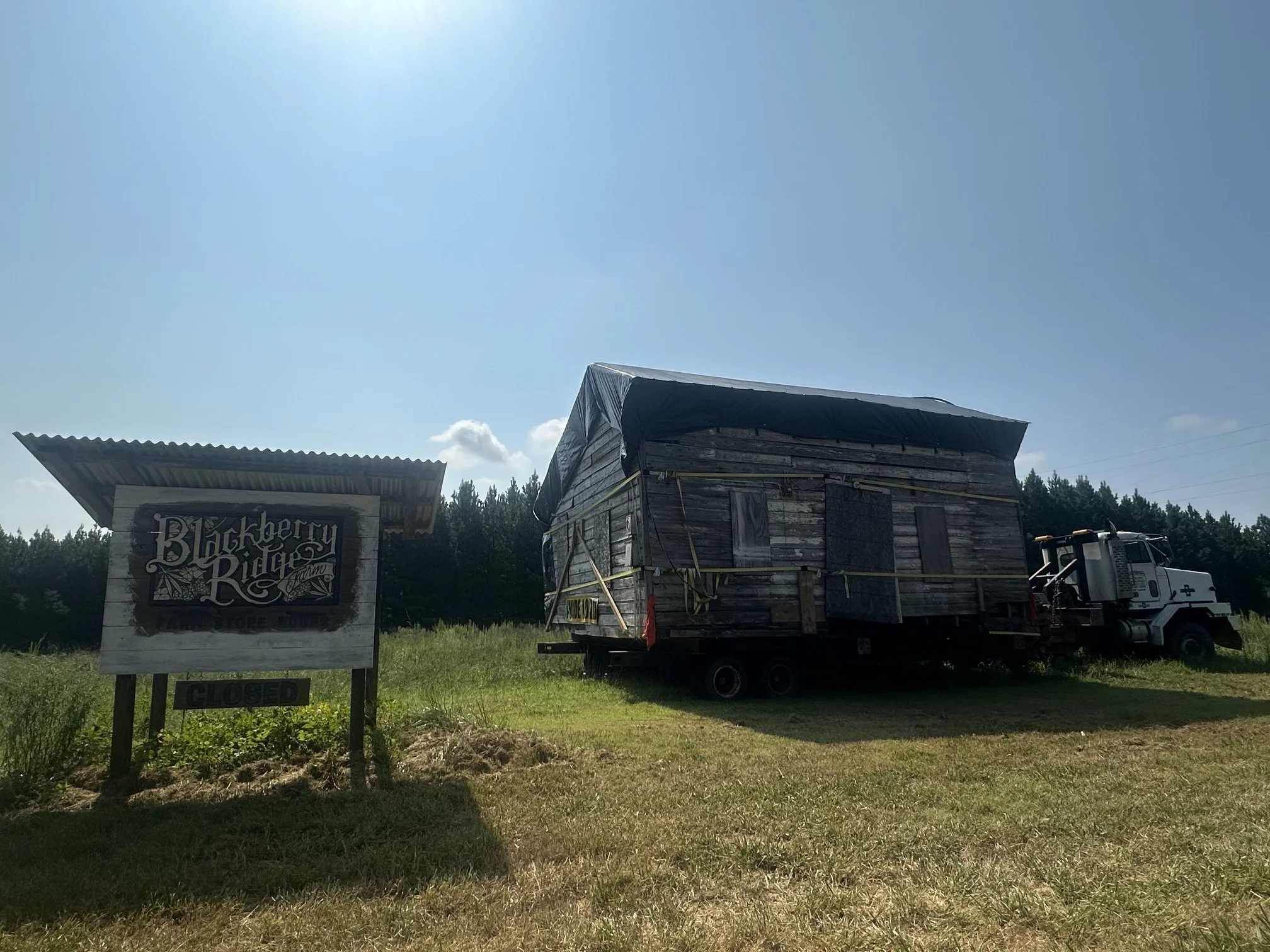 A wooden building with a sign that reads "Blackberry Ridge Farm" and a white truck parked in front. The building is enclosed with yellow caution tape, indicating it may be closed or under construction. There are trees and a clear blue sky in the background.