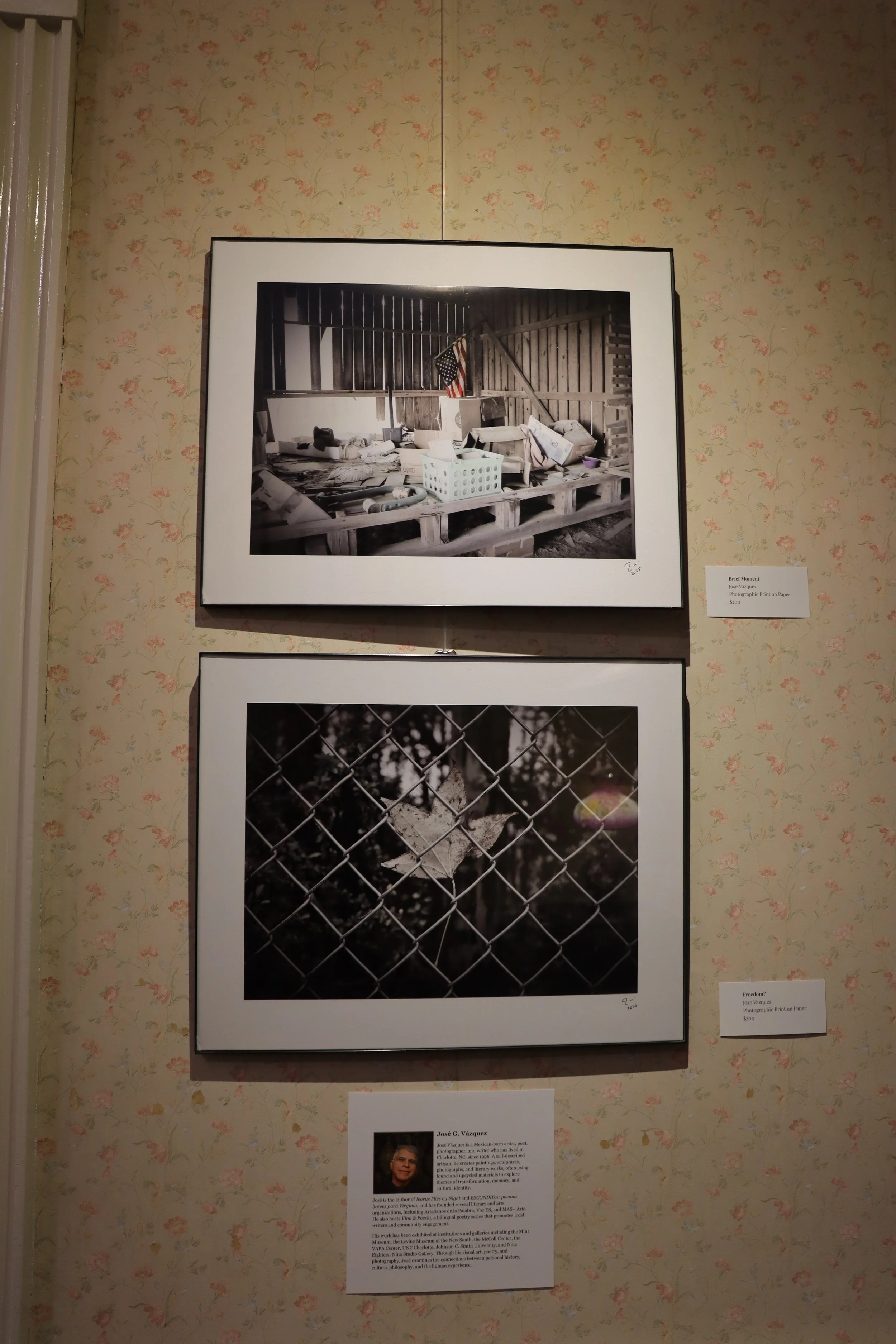 Two framed black-and-white photographs on a patterned wallpapered wall, with descriptions and an artist's biography next to them. The top photo depicts a cluttered attic space with furniture and an American flag, while the bottom photo shows a close-up of a chain-link fence with a tree and leaves in the background.