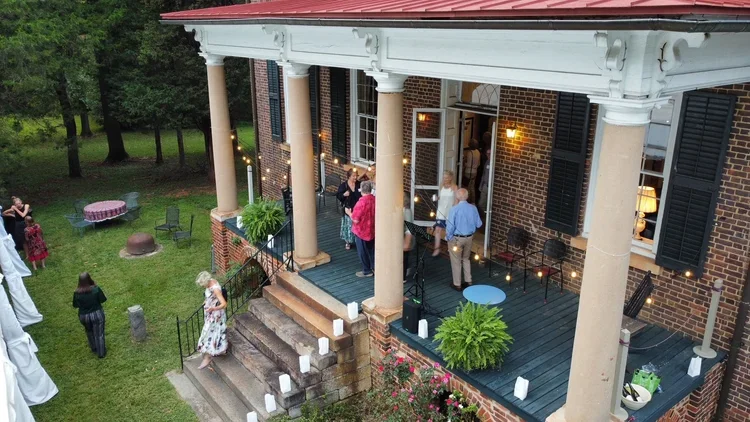 People gathering on the porch of a brick house with string lights, black shutters, and outdoor furniture, during a social event in the evening.