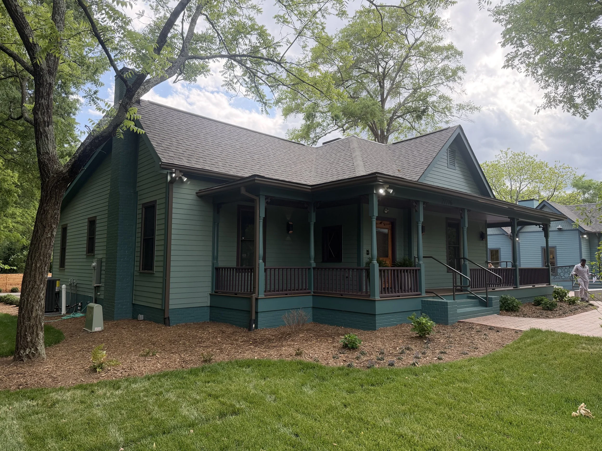 A house painted in light green with a porch, surrounded by trees and a well-kept lawn, under a partly cloudy sky.
