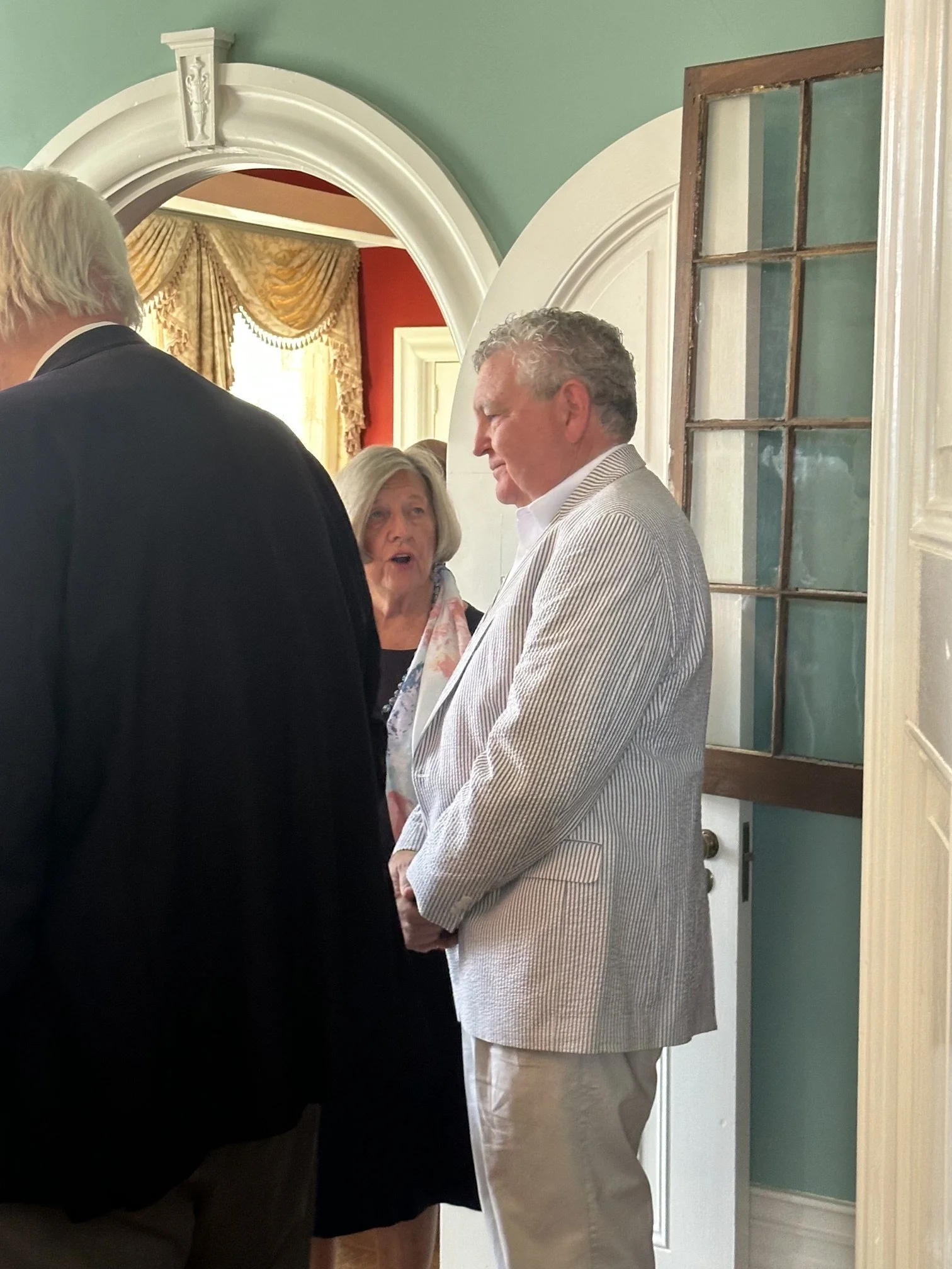 Three people in conversation inside a room with green walls, ornate white trims, a decorative window, and a gold curtain.